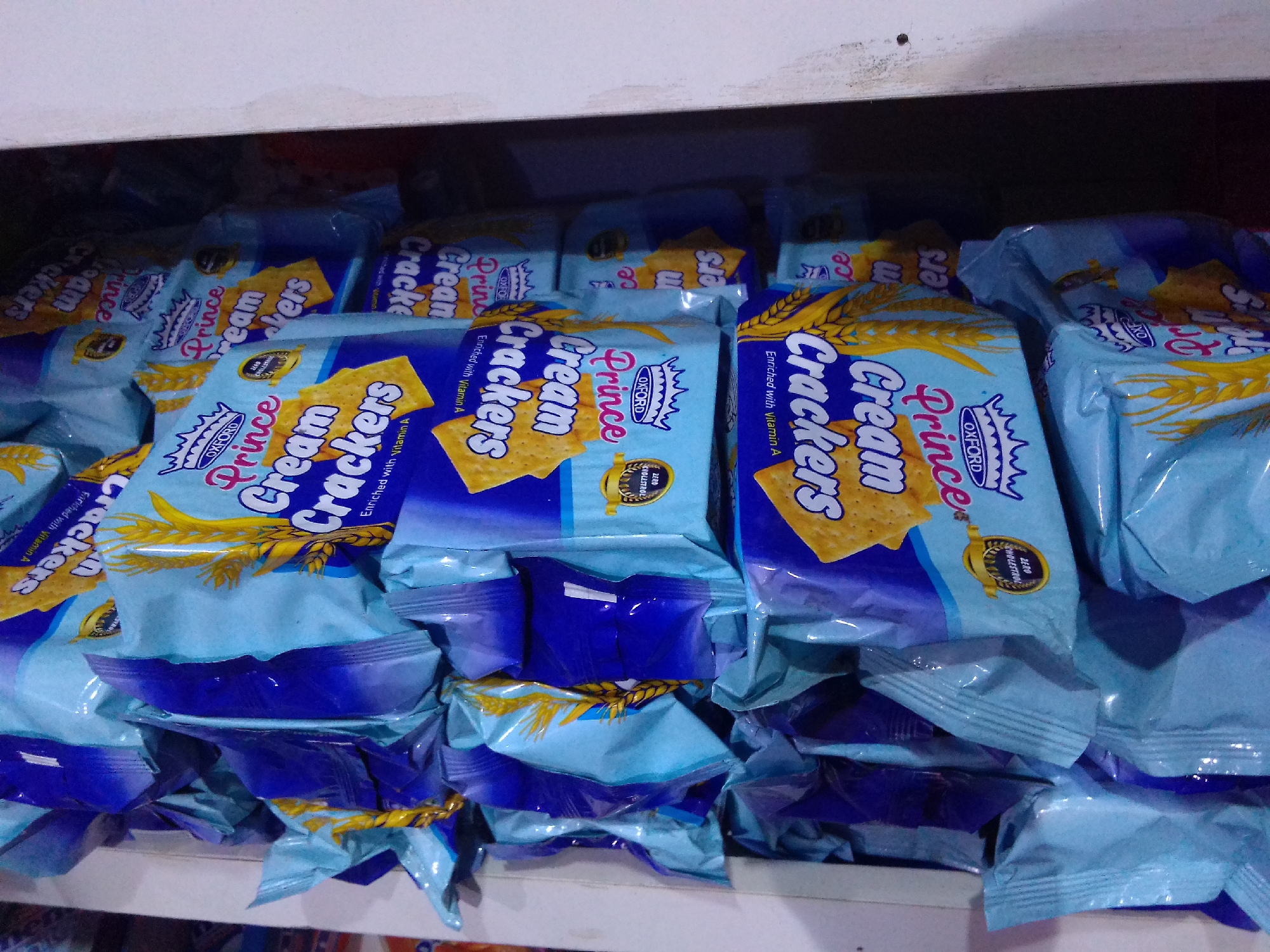 The image displays a close-up view of multiple packages of "Prince Cream Crackers" biscuits stacked on a shelf, likely in a retail setting. The packaging is predominantly light blue with prominent yellow and white text and imagery. The branding "Prince" is clearly visible in a stylized font, accompanied by the word "Cream Crackers." Some packages show an illustration of wheat stalks and a golden seal with text that appears to read "ZERO CHOLESTEROL." The lighting suggests an indoor environment, possibly a store or warehouse, with the items arranged in a slightly disorganized manner, typical of a display. The background is dark and out of focus, with the top edge of the shelf visible at the top of the frame. The overall impression is a visual inventory of a common consumer product. The location is described as Howayi, Nigeria, though this is not visually verifiable from the image content itself.