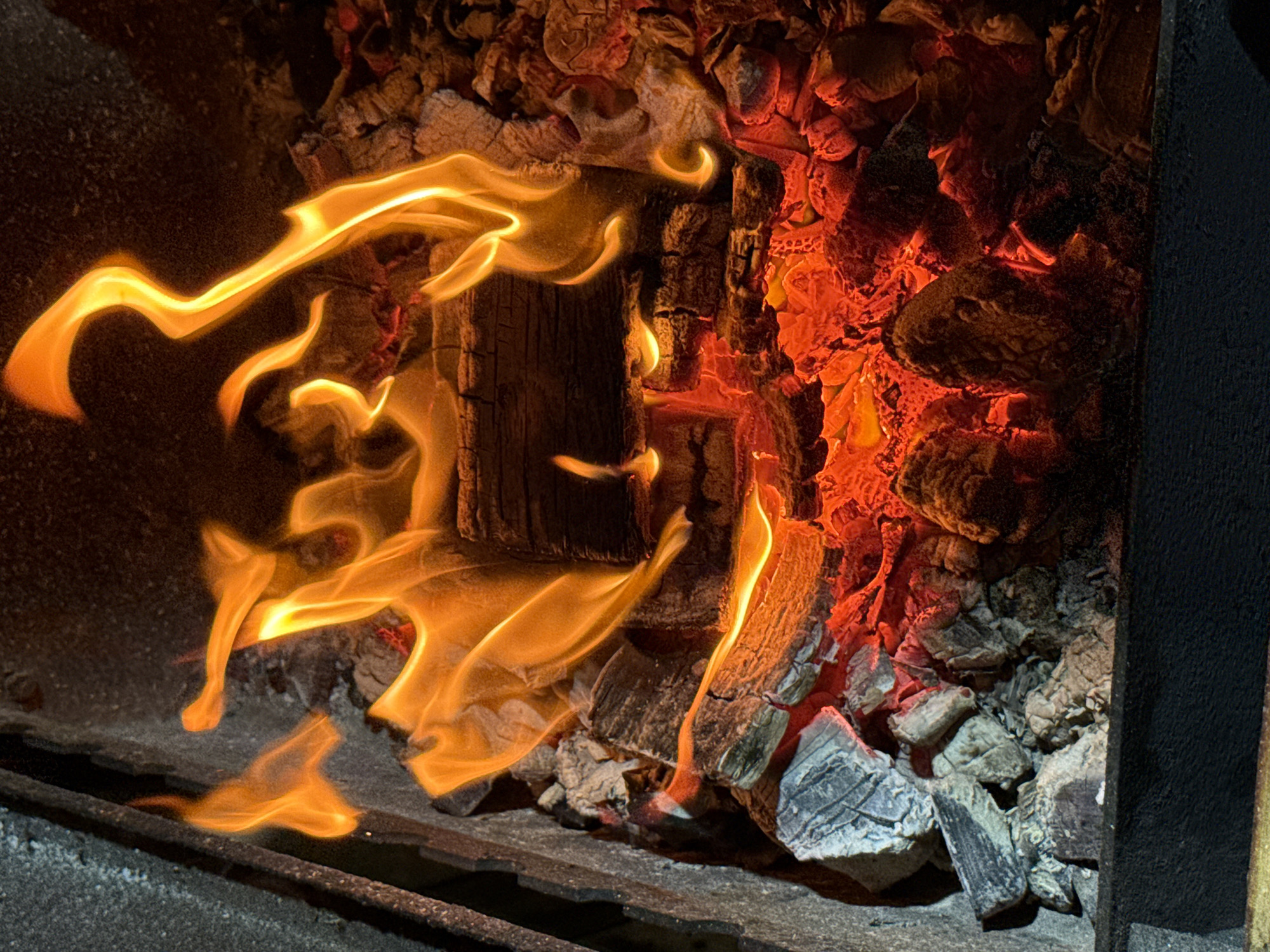 The image shows a close-up view of a fire burning in a fireplace or fire pit. There are logs of wood, burning embers, and flames that are orange and yellow. The background appears to be the textured stone or metal of the fireplace. The lighting is dim, with the fire providing the primary light source, casting a warm glow on the surrounding materials. There is no indication of the location in the city of Kamyshin, Russia, in the image itself.