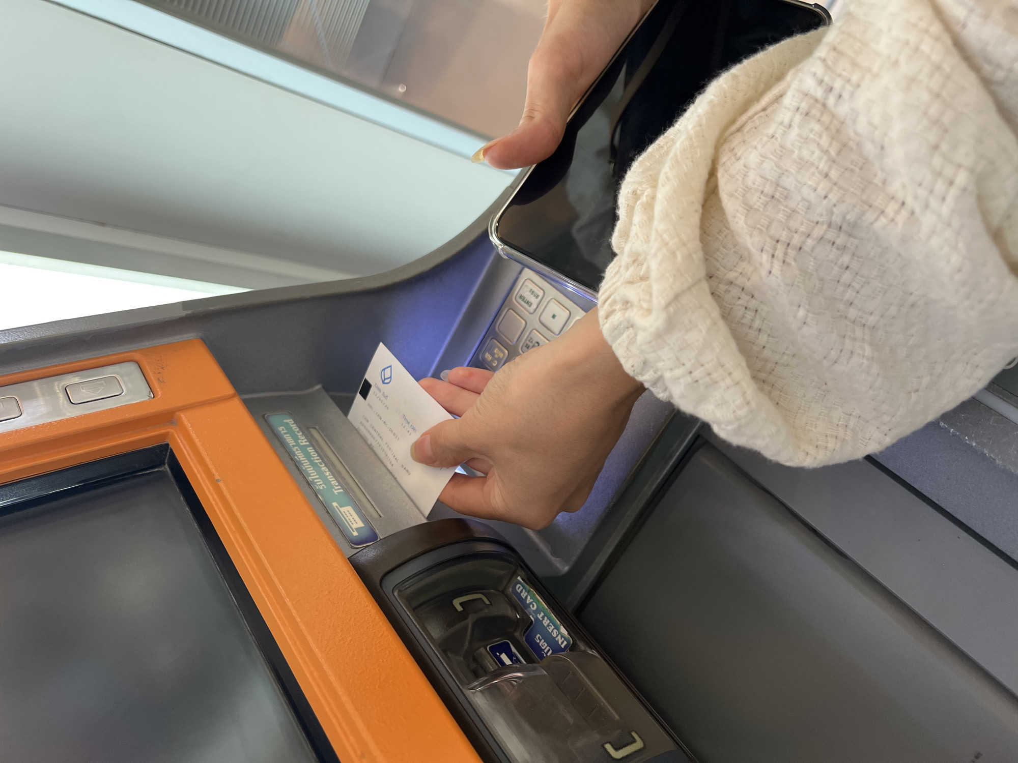 In the city of Ban Bang Rak, Thailand, a person is shown inserting a card into an ATM. The ATM has an orange and gray exterior. The screen of the ATM is visible and appears to be dark. A slot for inserting a card is clearly marked with the words "INSERT CARD" in English and Thai. The person's hands are visible, holding a white card and a black smartphone. The person is wearing a light-colored, textured sweater. The background shows a light-colored wall and a sliver of a ribbed surface. The scene captures a routine transaction at an automated teller machine.