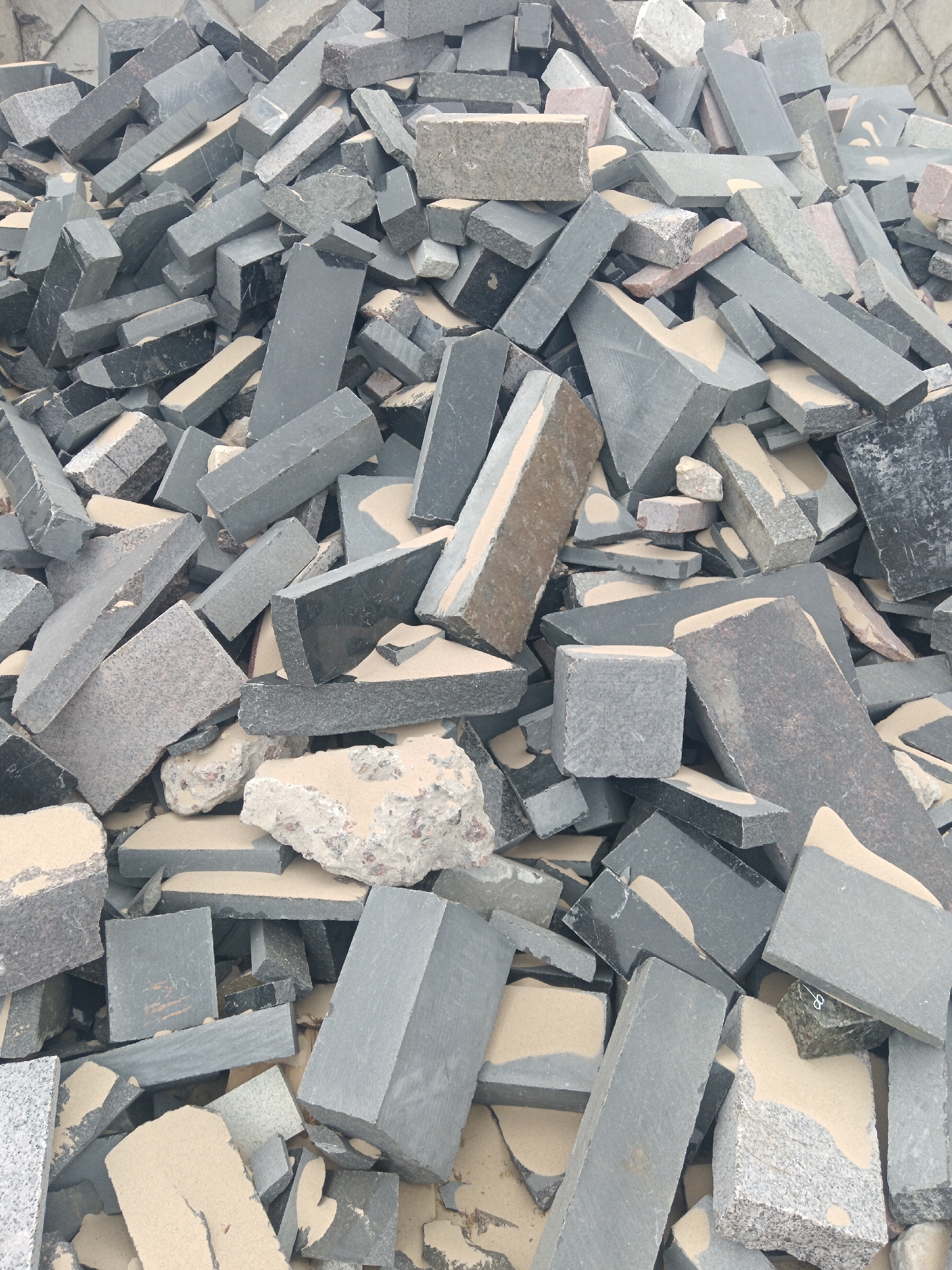 The image depicts a large pile of irregularly shaped and sized stone or concrete debris, possibly from a construction or demolition site. The color palette consists predominantly of gray, black, and beige hues. The debris includes rectangular, block-like pieces, with a mix of smooth and rough textures. Some pieces show internal fracturing or degradation. A light-colored, fine particulate matter, possibly sand or dust, covers much of the surface of the debris. The background shows a nondescript wall in a similar gray tone. The location, Яльгелево, Russia, cannot be determined from the visual information provided.