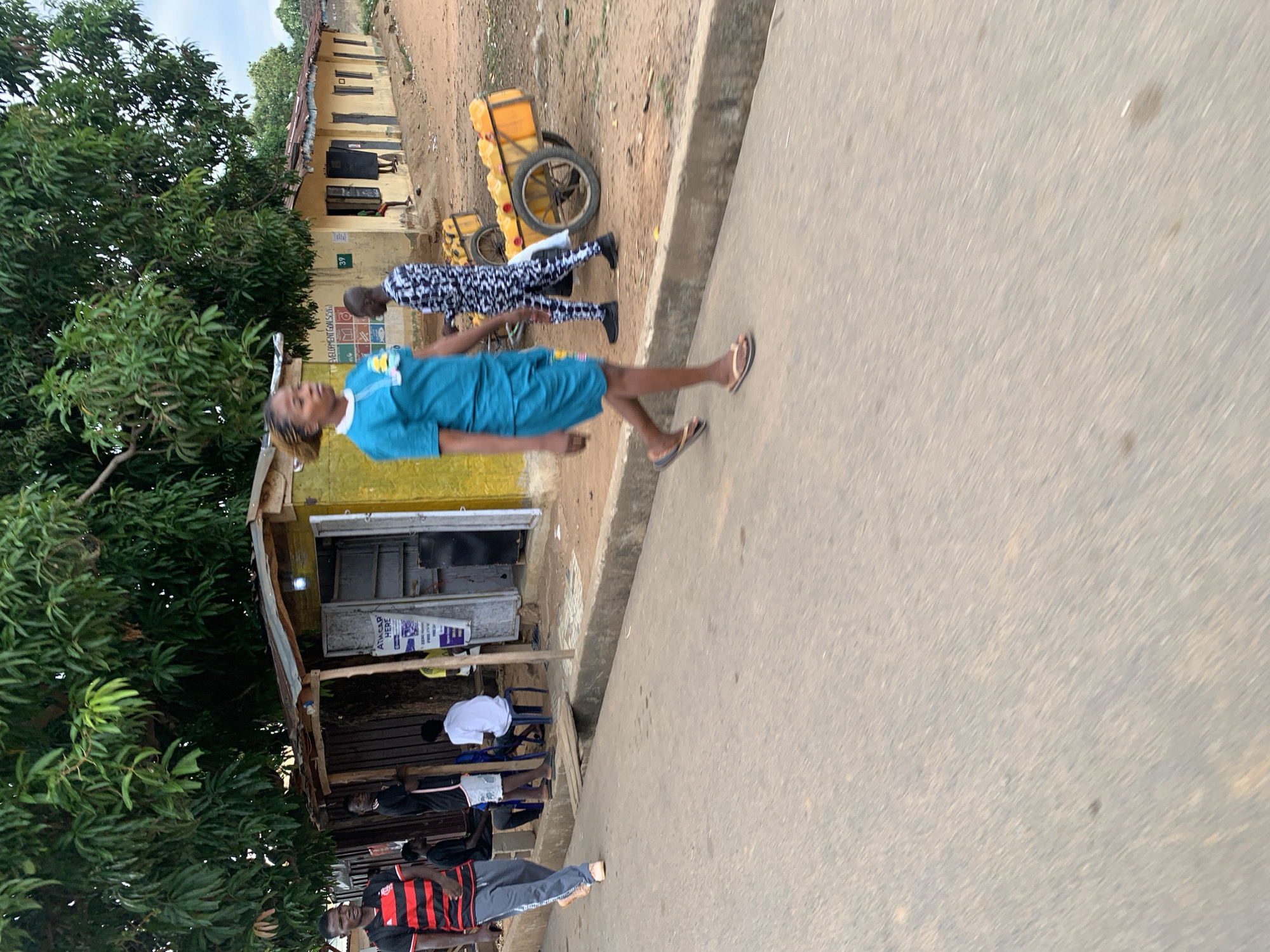 The image depicts a street scene in Akwanga, Nigeria. A woman in a blue dress and flip-flops is walking along the side of the road. Another person in a black and white patterned outfit is walking behind her, pulling a two-wheeled cart loaded with yellow containers. Buildings line the left side of the street, shaded by trees. People are sitting outside one of the buildings. The road surface is concrete.