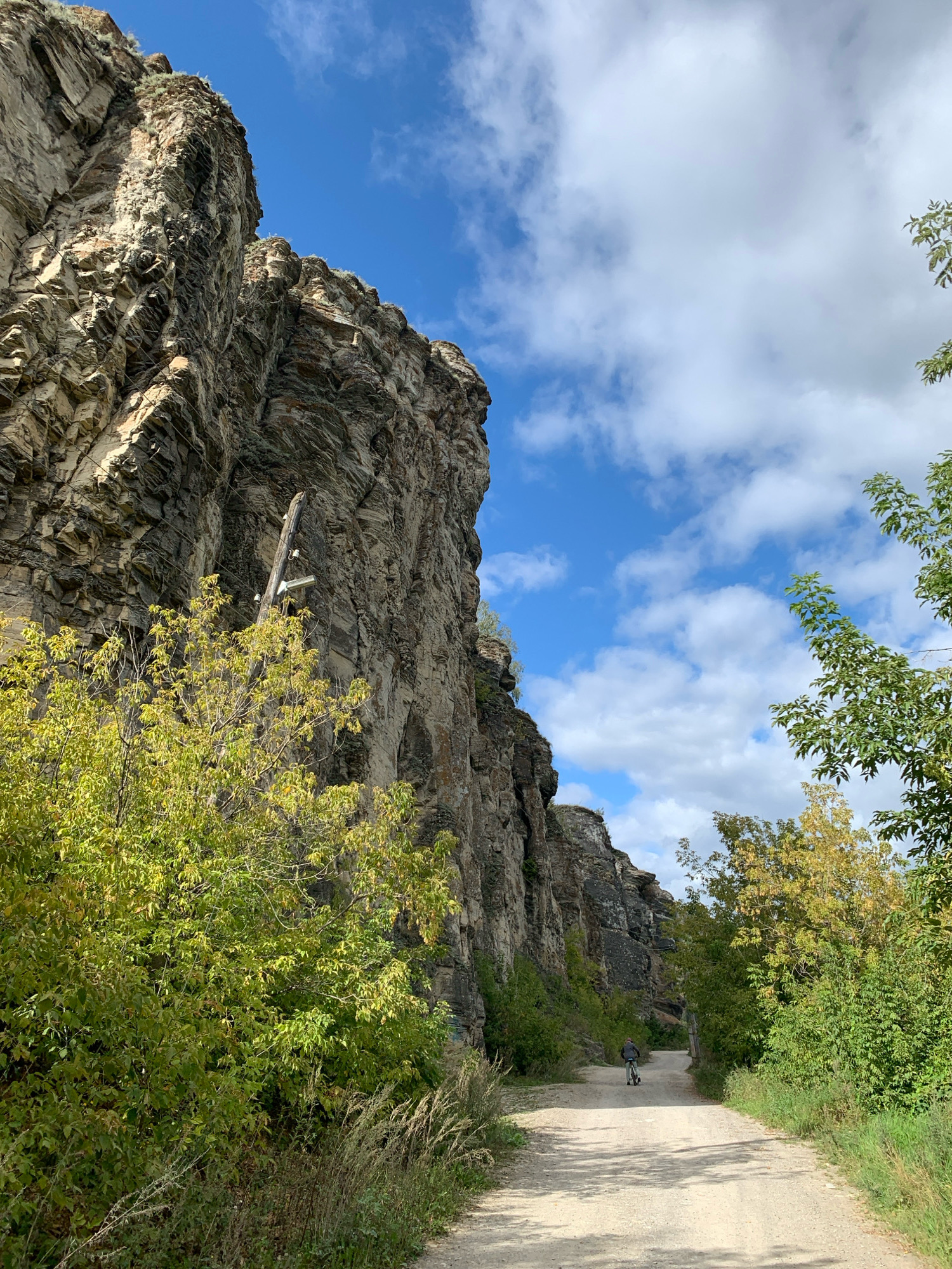 The image depicts a dirt path leading through a rugged landscape in the city of Minyar, Russia. On the left, a towering cliff face, composed of stratified rock, dominates the scene. Green foliage and dry grasses grow at the base of the cliff and along the path. On the right, the path is bordered by lush green trees, some of which show signs of autumnal color change. In the distance, a person is seen riding a bicycle on the path. The sky above is a mixture of blue with scattered white clouds. The overall impression is of a natural, somewhat remote setting within the city.