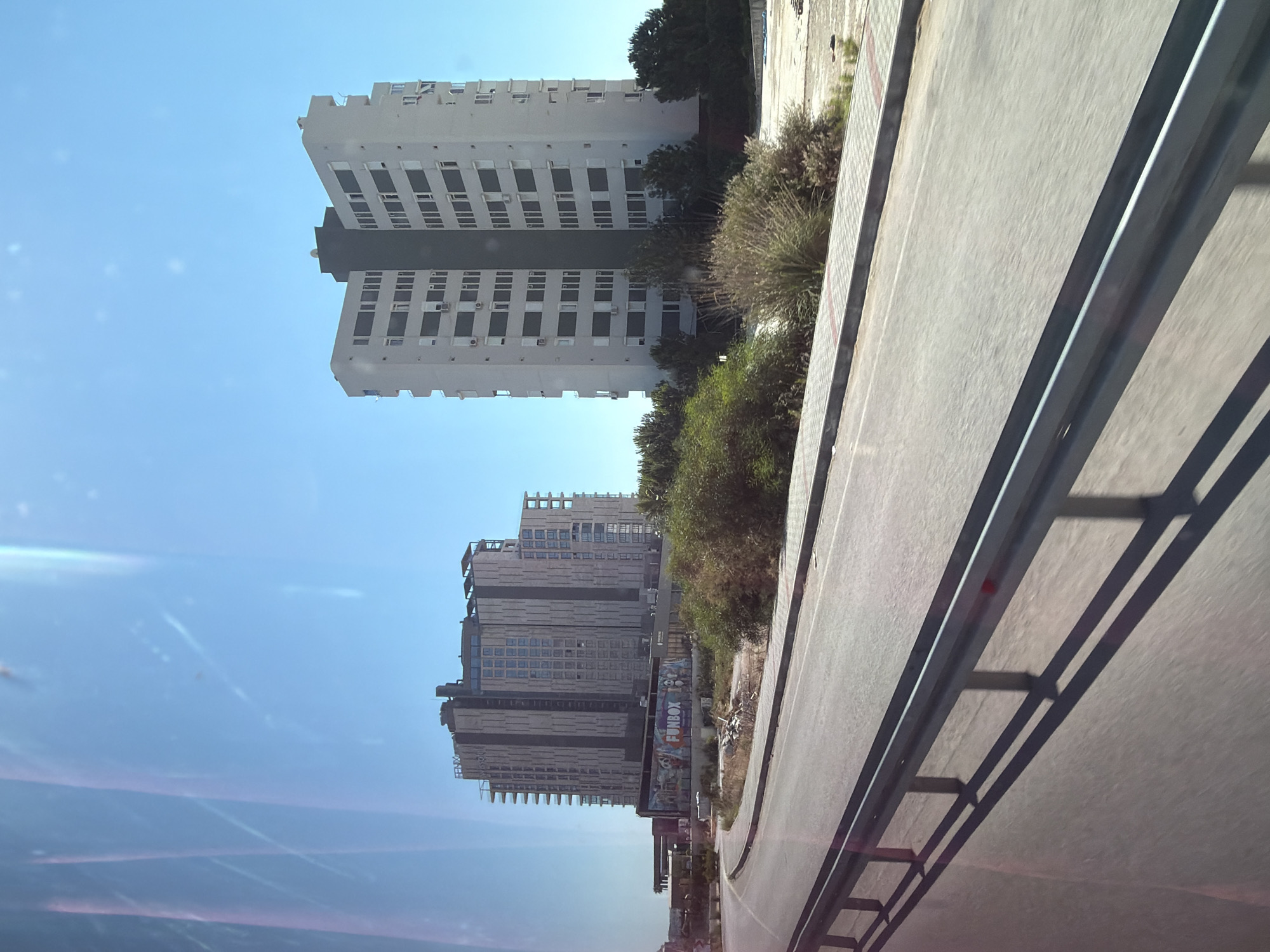 The image captures a sunny day in Konyaaltı, Turkey, with a clear blue sky. In the foreground, a road with a metal guardrail runs diagonally from the bottom left to the bottom right. The road appears to be in motion, suggested by the slight blur. Beyond the road, a large, modern building complex dominates the scene. The building on the left is a tall, multi-story structure with a facade of vertical gray panels and windows, appearing to be under construction or recently completed. A large advertisement for "FUNBOX" is visible on a lower section of this building. To the right, a taller, white building with a dark central core stands out. This building also features numerous windows and air conditioning units. Lush green trees and dry brush surround the base of the buildings. The overall scene depicts an urban environment with modern architecture juxtaposed with natural elements.