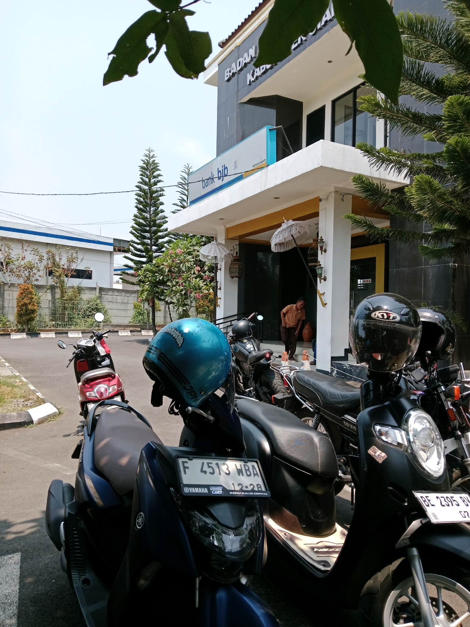In Karangtengah, Indonesia, a clear, bright day is captured in front of a Bank BJB building. The building's facade features a dark gray exterior with the Bank BJB logo prominently displayed. A white awning extends from the building, over a yellow-accented entrance. Several motorcycles are parked in front of the building, including a blue Yamaha with license plate F 4513 WBA and a black motorcycle with license plate BE 2395 BY. A man in a brown shirt is walking out of the building's entrance, carrying what appears to be a large clay pot. Green foliage from nearby trees frames the top and right side of the image. The ground is paved, with a white-lined parking area visible.
