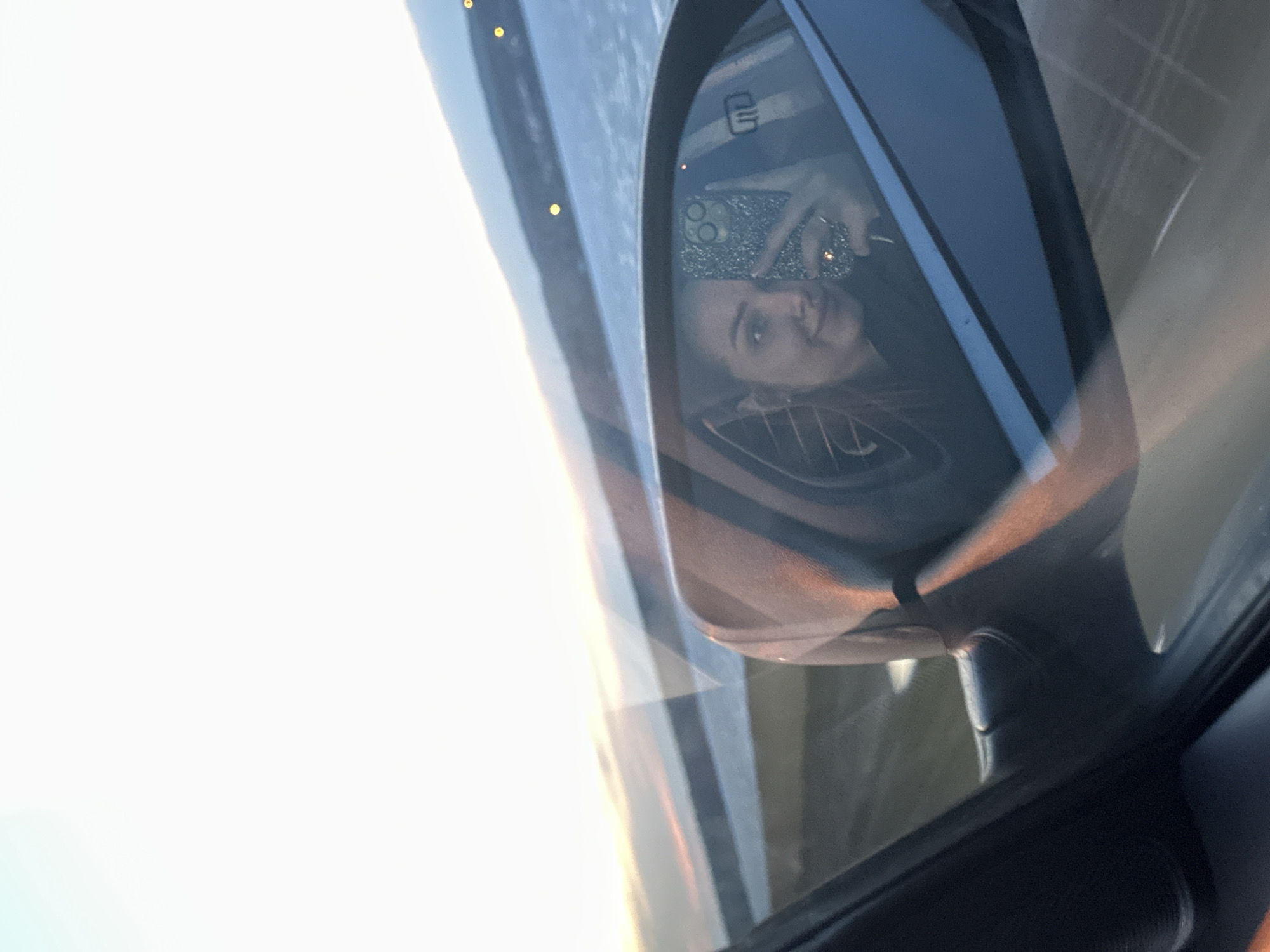 A selfie reflection in a car side mirror depicts a woman holding a phone with a glittery case. The background visible through the mirror shows a body of water and shoreline. The exact location in Chaikovsky, Russia, cannot be determined from the image alone.