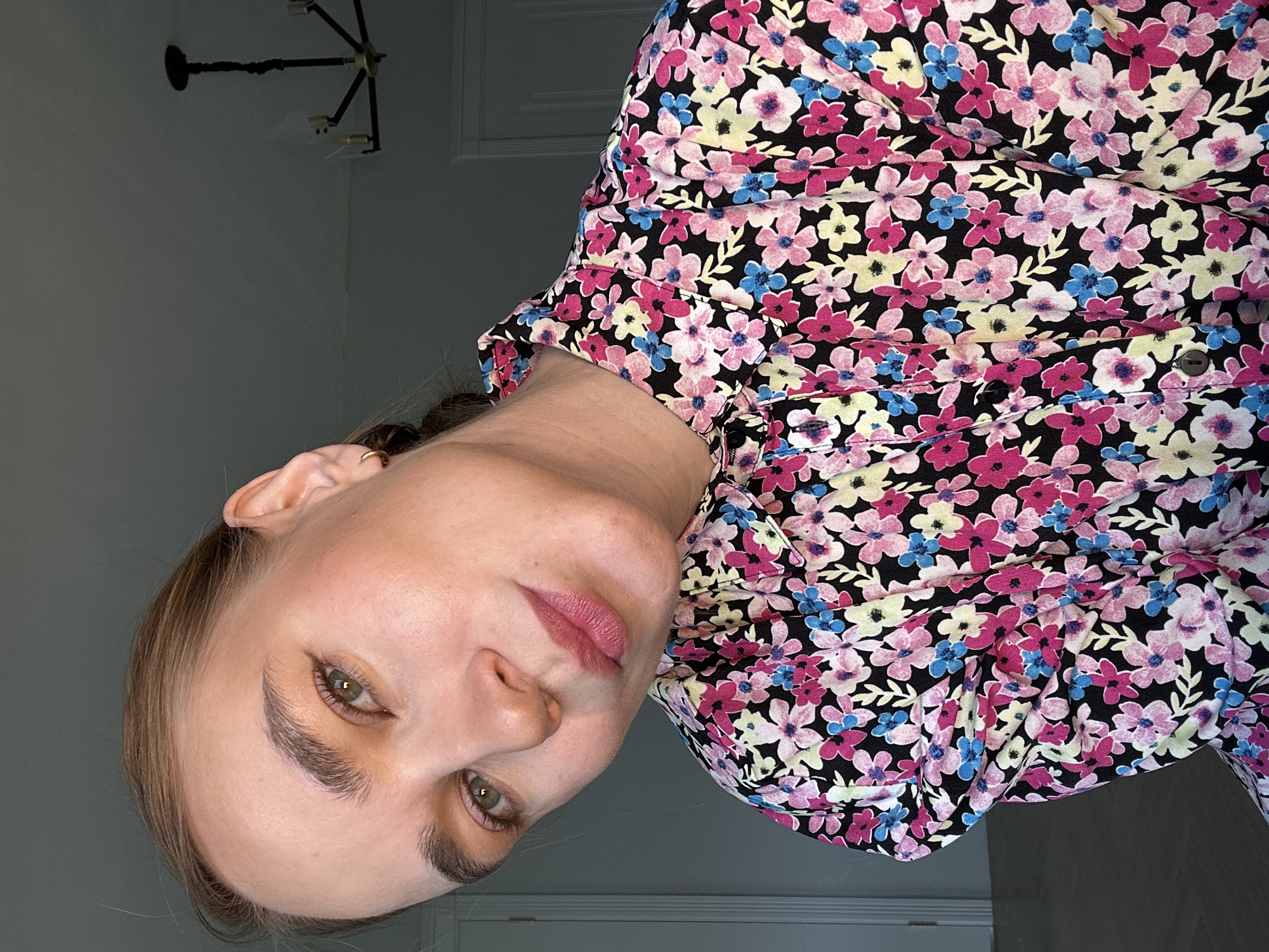 A woman is facing the camera, rotated counter-clockwise. She has fair skin, light brown hair pulled back, and green eyes. She is wearing a floral print shirt with a black background and pink, light yellow, and blue flowers. The background is a light gray wall with a dark chandelier visible in the upper left and a white door in the back. There is no indication of location.