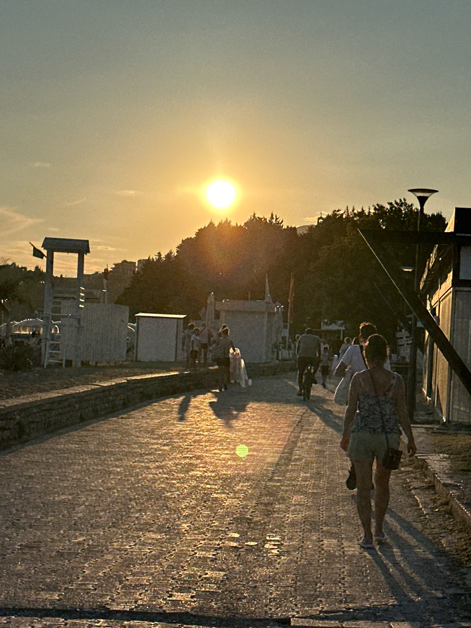 The image depicts a sunset scene on a paved walkway, likely in the city of Boreti, Montenegro, given the context. A woman in a patterned top and shorts is walking away from the camera, casting a long shadow on the ground. Several other individuals are visible in the distance, some walking and one riding a bicycle. The setting sun is prominent in the upper portion of the image, casting a warm glow and creating reflections on the wet-looking pavement. In the background, there are trees and buildings, including a structure resembling a lifeguard tower. The overall atmosphere is serene, with a sense of people enjoying the evening.