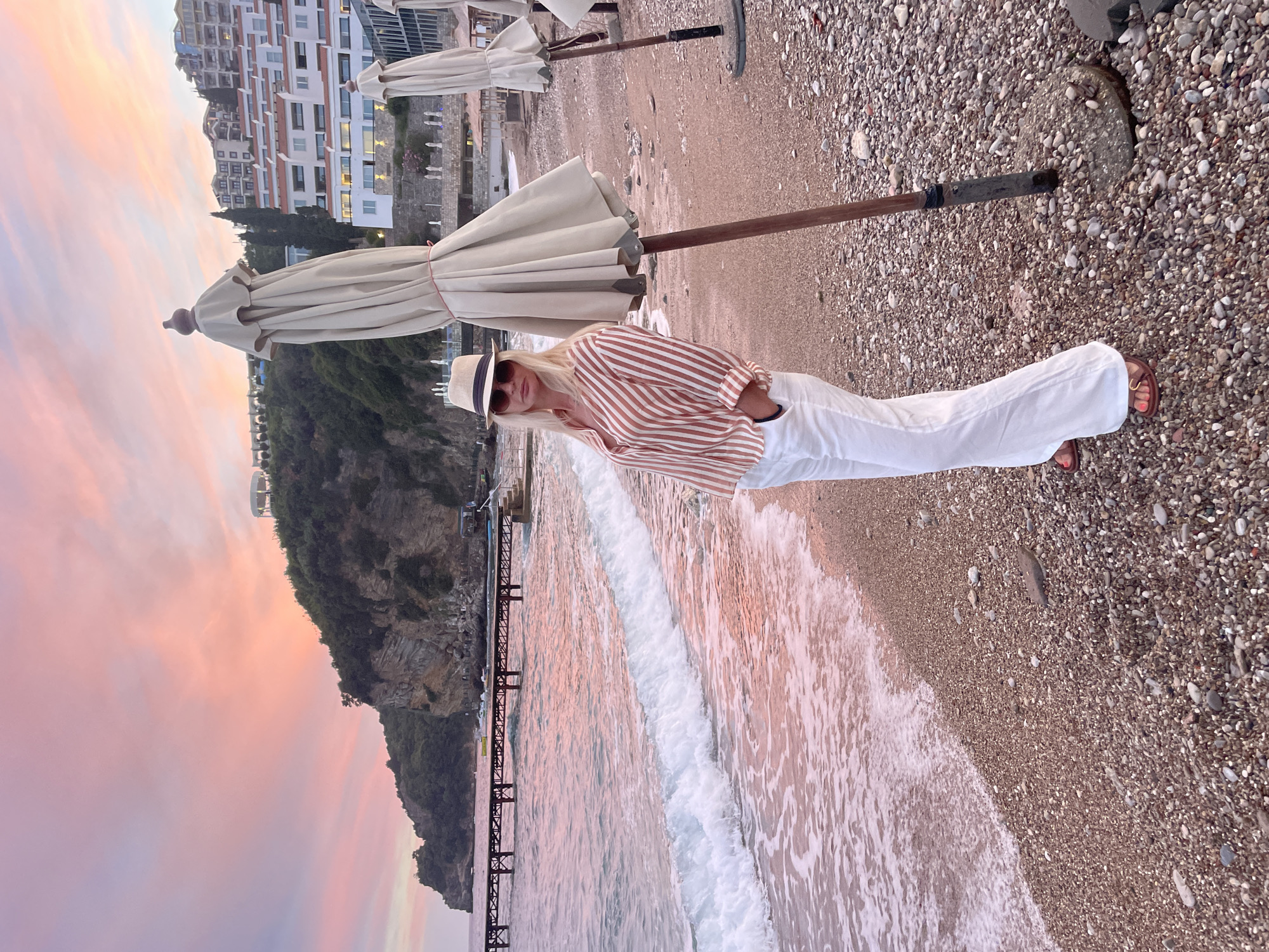 A woman stands on a pebbled beach in Budva, Montenegro, facing the camera. She is wearing a straw hat, sunglasses, a striped shirt, and white wide-leg pants. Her hands are in her pockets. Behind her, the ocean meets the shore with small waves. In the background, a pier extends into the water, and a hillside is covered with lush green vegetation and buildings. The sky is painted with soft pink and purple hues of a sunset. Several closed beach umbrellas are visible on the right side of the image.