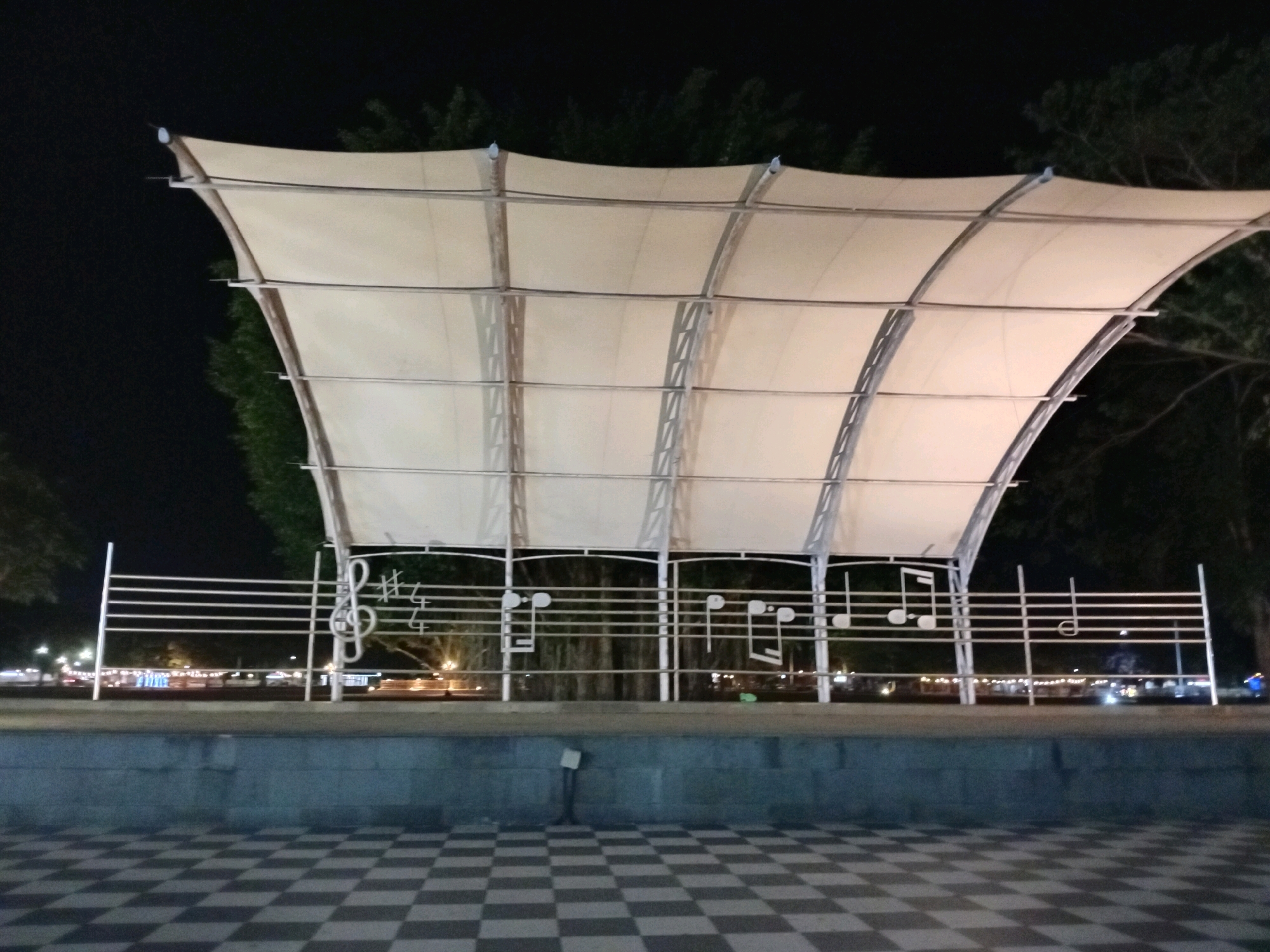 The image shows an outdoor stage at night in Purworejo, Indonesia. The stage has a large, curved white canopy supported by a metal frame. In front of the stage, there is a decorative metal railing designed to resemble a musical staff, complete with treble clef and musical notes. The foreground features a checkered pavement. In the background, trees and illuminated buildings are visible, suggesting an urban environment. The lighting suggests it is a public space, possibly a park or plaza.