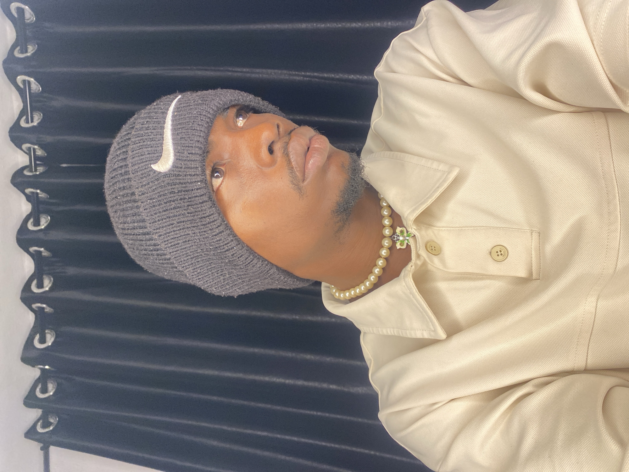In a medium close-up shot, a Black individual with light brown skin wears a grey knit beanie with a white Nike logo on the front, a pearl necklace with a green pendant, and a cream-colored collared shirt. The individual is looking off to the right of the frame. Behind them, a dark grey curtain with silver grommets is visible. The image appears to be taken indoors, possibly in a residential setting. The lighting suggests it could be artificial light. The setting is not identifiable as a specific location in Abakaliki, Nigeria.