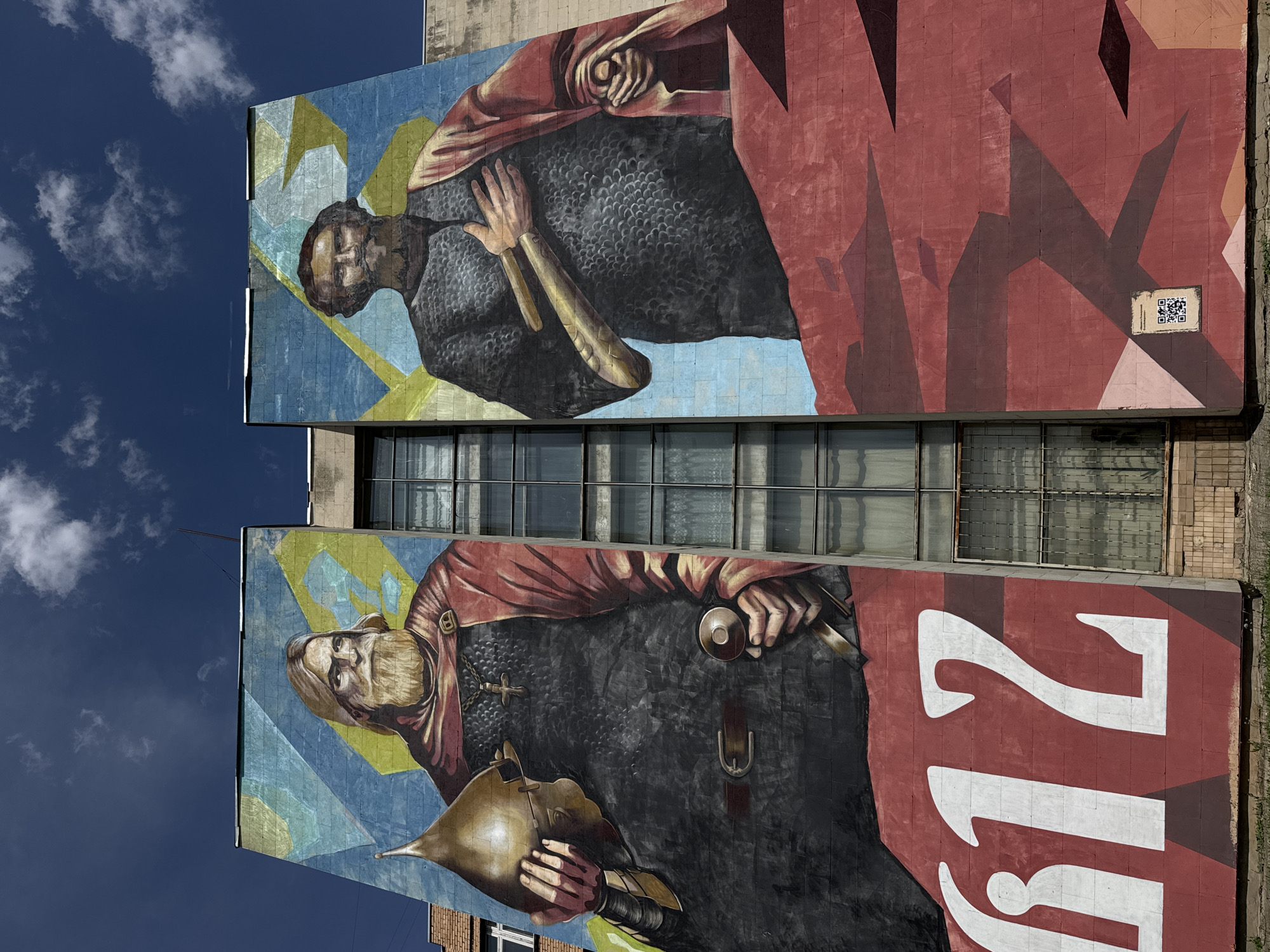 Here is a detailed description of the image:
The image shows a large mural painted on the side of a building in an urban environment. The mural depicts two figures in historical attire, likely referencing a historical event or figure. The bottom portion of the mural features a figure with a beard, wearing what appears to be chainmail and a red cloak. He holds a helmet in one hand and a sword in the other. Beside him, large white numbers "212" are painted on a red background. Above this, there is another section of the mural depicting a second figure, also in historical clothing, with a similar red cloak. The figures are set against a background of blue and yellow geometric shapes. The mural covers a significant portion of the building's facade. The building itself appears to be a multi-story structure with rows of windows between the two sections of the mural. A QR code is visible in the top right corner of the mural. The sky is visible behind the building and mural, showing a clear blue sky with some clouds.