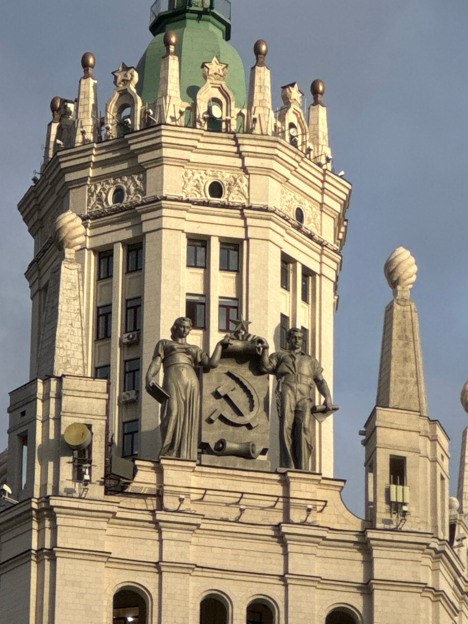 The image captures the upper section of the Kotelnicheskaya Embankment Building, a Stalinist skyscraper located in Moscow, Russia. Prominently featured are two statues positioned above the building's cornice. The statue on the left depicts a woman in a flowing robe, holding a book. To her right, a statue represents a man in work clothes, holding a scroll. Between them, a large Soviet emblem, the hammer and sickle, is displayed on a shield. The building itself is constructed of light-colored stone and features numerous windows. The sky above is clear and blue.