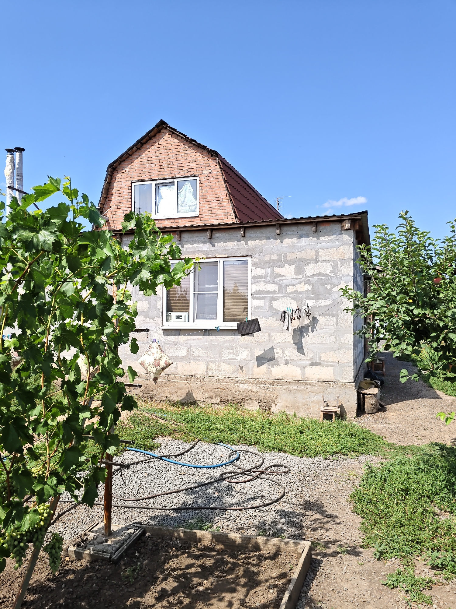 The image depicts a rural scene with a two-story house under a clear blue sky. The lower portion of the house is constructed from cinder blocks, while the upper, attic section is brick. Two windows are visible on the lower level, and one on the upper level. Laundry, including what appears to be a pillow, is hung on a clothesline attached to the side of the house. To the left of the house, a grape vine with green grapes is growing. To the right, an apple tree with some fruit is laden with green leaves. The ground around the house is a mixture of gravel and dirt, with patches of green grass. A hose lies coiled on the gravel. Several rustic outdoor items, including a wooden stool and a tree stump, are present in the yard. The scene is situated in the city of Nizhnedonskoy, Russia.