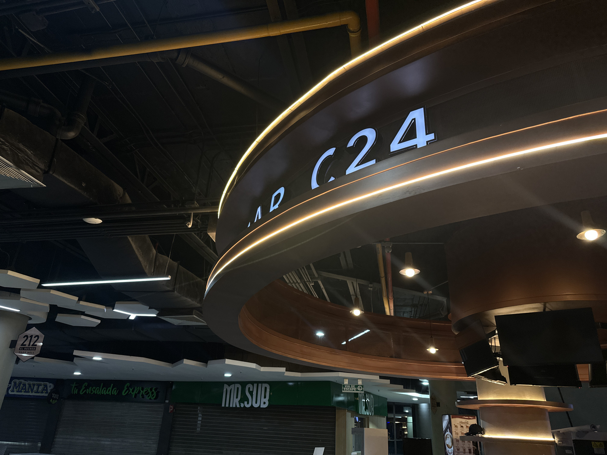 The image captures an interior view of El Recreo Food Hall, located in Caracas, Venezuela. A prominent circular overhead sign displays the text "FOOD C24" in illuminated white letters, framed by warm LED strip lights. The background reveals the food hall's layout with closed storefronts, including "MANIA to Ensalada Express" and "MR. SUB". The food hall's ceiling features geometric white panels and visible infrastructure, such as pipes and conduit. The light from the LED lights illuminates the ceiling. The color scheme is predominantly dark with contrast. There are several displays of information on screens.