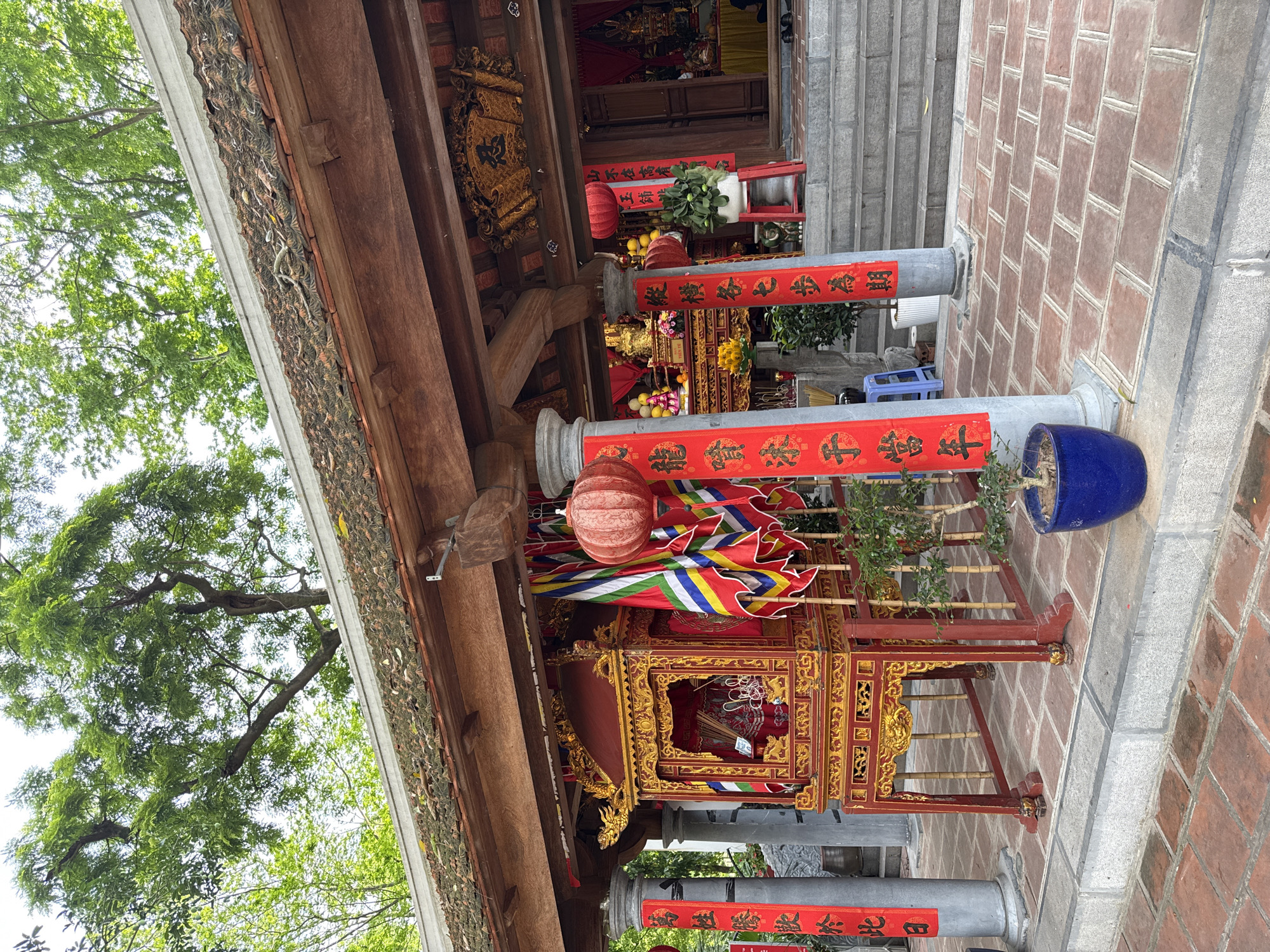 The image shows a traditional Vietnamese temple scene, likely located in Hà Nội, Vietnam. A red and gold palanquin is prominently displayed, adorned with colorful banners and a spherical lantern. Two columns are draped with red banners bearing Chinese characters. The temple structure features a tiled roof and wooden beams. In the background, a statue is visible on an altar. Brick paving is underfoot. A potted plant sits to the side.