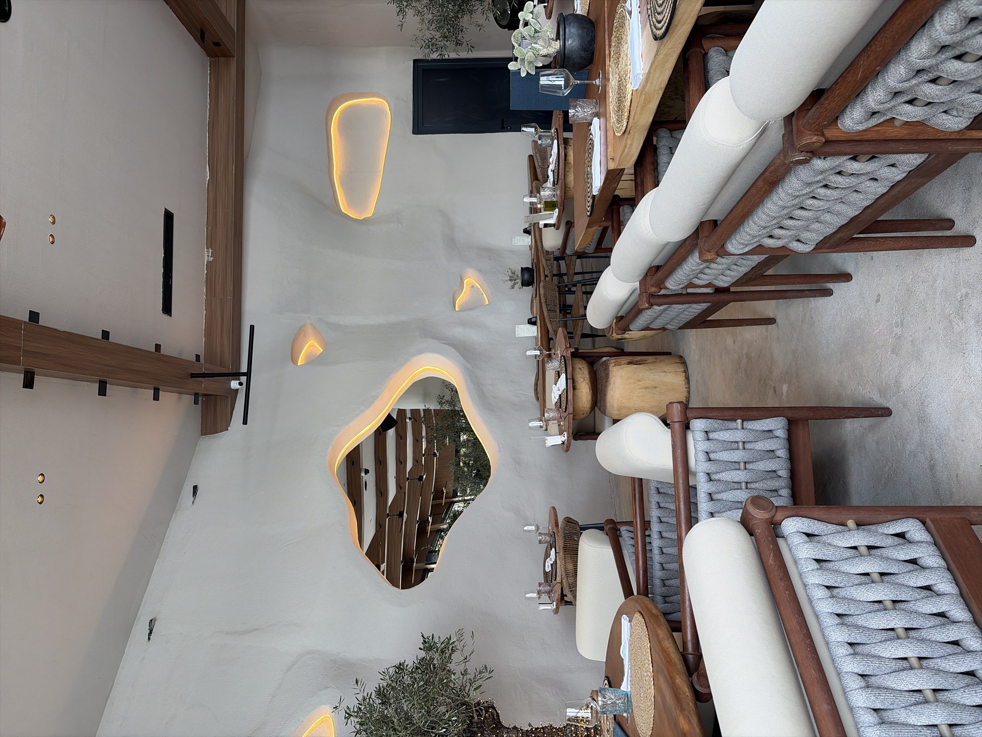 This image depicts the interior of a modern, upscale restaurant or cafe located in the city of Unknown, Unknown, presented from a slightly elevated perspective looking into the dining area. No individuals are present in the scene.
The environment features textured, off-white plaster walls with organic, undulated shapes. Several of these shapes are recessed and internally illuminated with a warm orange glow. A prominent, irregularly shaped mirror, also outlined with a warm orange light, is centrally positioned on the left wall, reflecting an overhead ceiling with wooden beams and what appears to be an exterior or another section of the restaurant through its opening. The ceiling itself is flat and white, interspersed with dark wooden beams. Recessed spotlights are visible in the ceiling, along with a rectangular black air vent and a small black security camera mounted on a beam.
The flooring is a uniform light gray, possibly concrete or polished stone.
The dining area contains numerous tables and chairs, meticulously set for service. Tables are primarily constructed from natural wood, exhibiting varying shapes and sizes; some are round, others rectangular, with visible wood grain and sometimes rustic, uneven edges. Several tables feature thick, cylindrical or block-like wooden bases. Seating comprises two main styles: dark brown woven rattan or wicker chairs, and more contemporary armchairs with dark wood frames, gray woven rope backs and sides, and light cream-colored upholstered seat and back cushions. These armchairs are often arranged in pairs or longer bench-like configurations.
Each table is set with a woven placemat (either round tan or rectangular dark-patterned), a folded white napkin, a clear wine glass, a clear water glass (some with a blue textured pattern), and small condiment bottles. Many tables also feature small potted plants, including cacti and succulents, or small olive tree saplings. Larger olive trees, one adorned with small string lights, flank the dining space. In the background, a dark, minimalist door is visible.