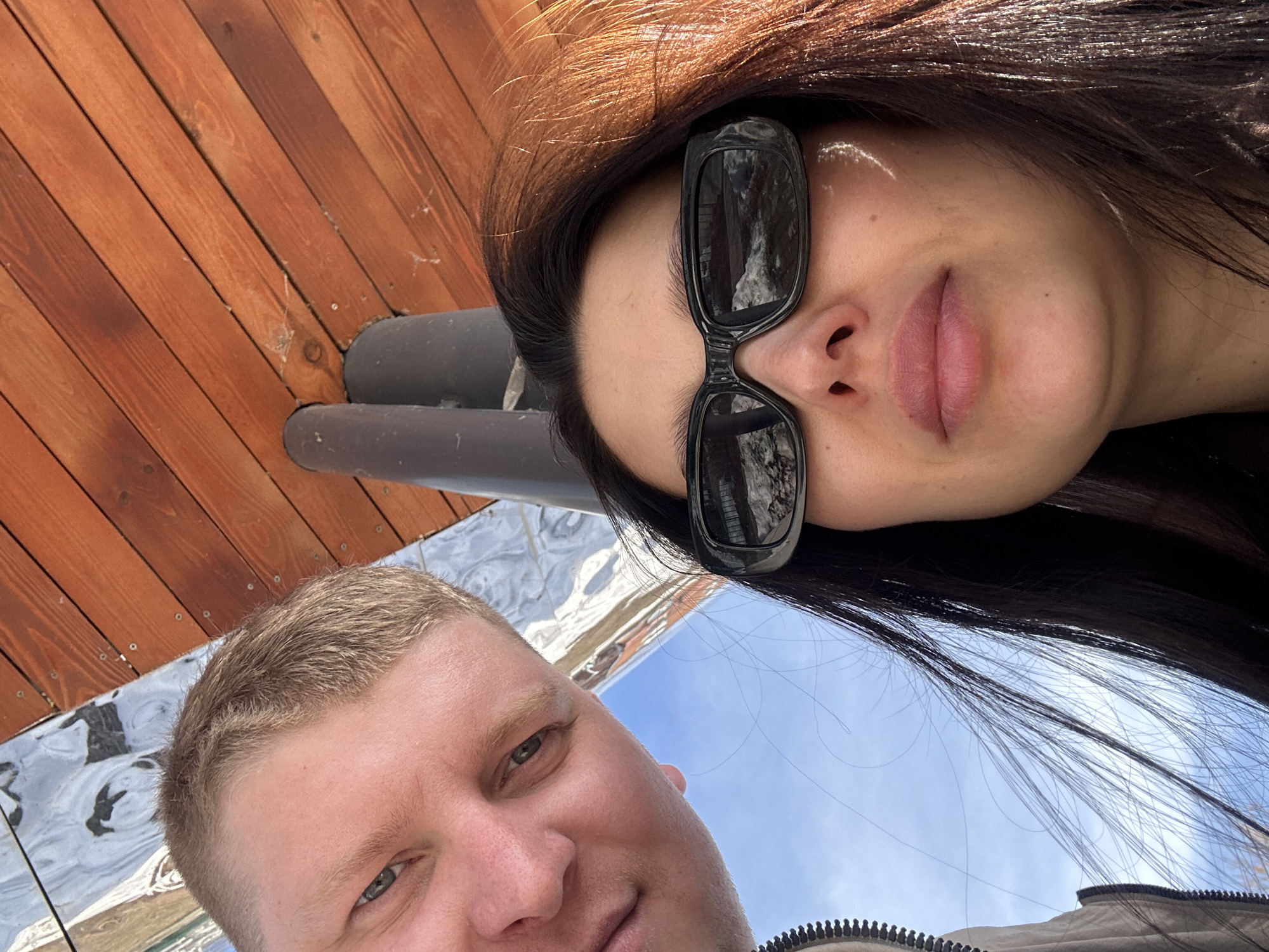 The image is a selfie taken by two people outdoors. A woman with long, dark hair and sunglasses is positioned at the top of the frame, while a man with short, blond hair is at the bottom. They are likely holding the camera together, and the perspective is angled upwards towards the sky. The background reveals some elements, including a wooden structure with pipes, some reflective panels, and a patch of blue sky. The environment suggests an outdoor location, possibly in a city. Given the request for the location Votkinsk, Russia, it can be inferred that this photograph was likely taken in that city.