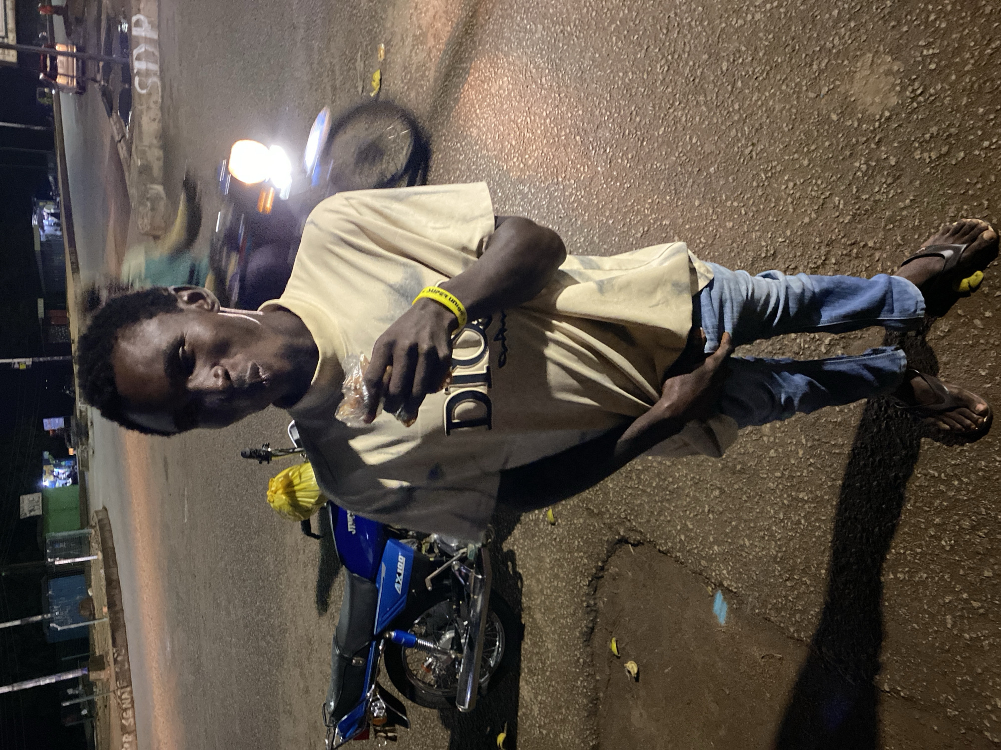 A young Black male, with short dark hair and dark skin, stands in the middle of a paved street at night in Bukuru, Nigeria. He is wearing a light tan crewneck t-shirt with the word "DIOR" printed on the front in large, dark letters, light blue denim jeans, and dark flip-flops. A yellow wristband is visible on his right wrist. He is holding a small, light brown food item to his mouth and appears to be eating it.
To his left, a blue motorcycle with black seats is parked, facing right, with a yellow plastic bag tied to the back. The motorcycle has "AX 100" printed on its side. In the background, there are streetlights casting illumination on the road and sidewalk. Several structures, possibly shops or buildings, are visible behind the street. A vehicle with illuminated headlights is blurred in motion on the road in the upper right portion of the image. The ground is paved and appears slightly wet in some areas. The overall lighting suggests it is nighttime.