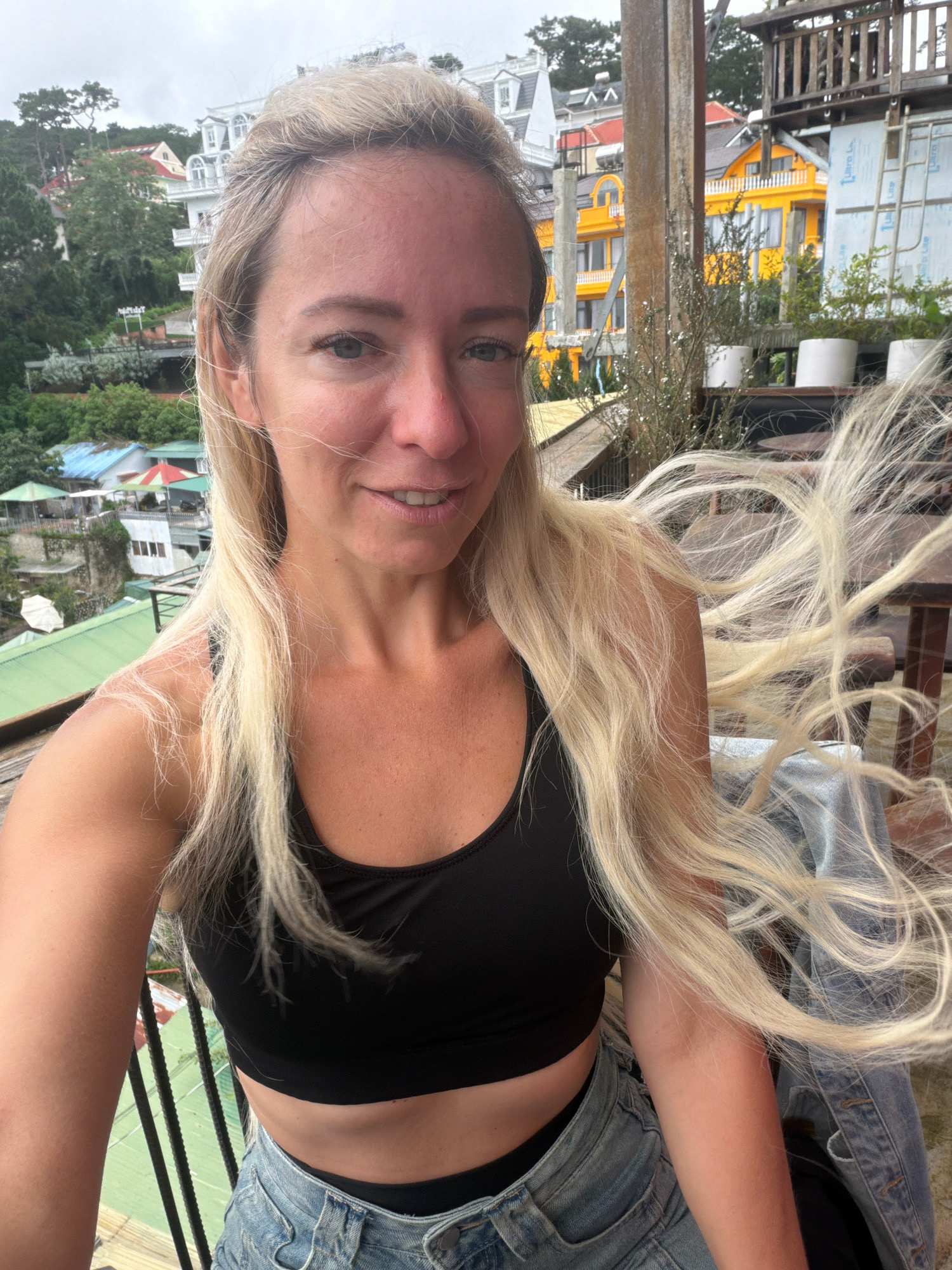 A young Caucasian woman with long blonde hair stands on a balcony overlooking a hillside town. The woman is wearing a black crop top and denim shorts. Her hair is blowing in the wind. In the background, there are colorful buildings, green trees, and a cloudy sky. The style of the buildings suggests a European influence, consistent with the French colonial architecture found in Da Lat, Vietnam. Specifically, the multi-story buildings with white trim and steep roofs are characteristic of villas in Da Lat. The woman is taking a selfie with her phone.