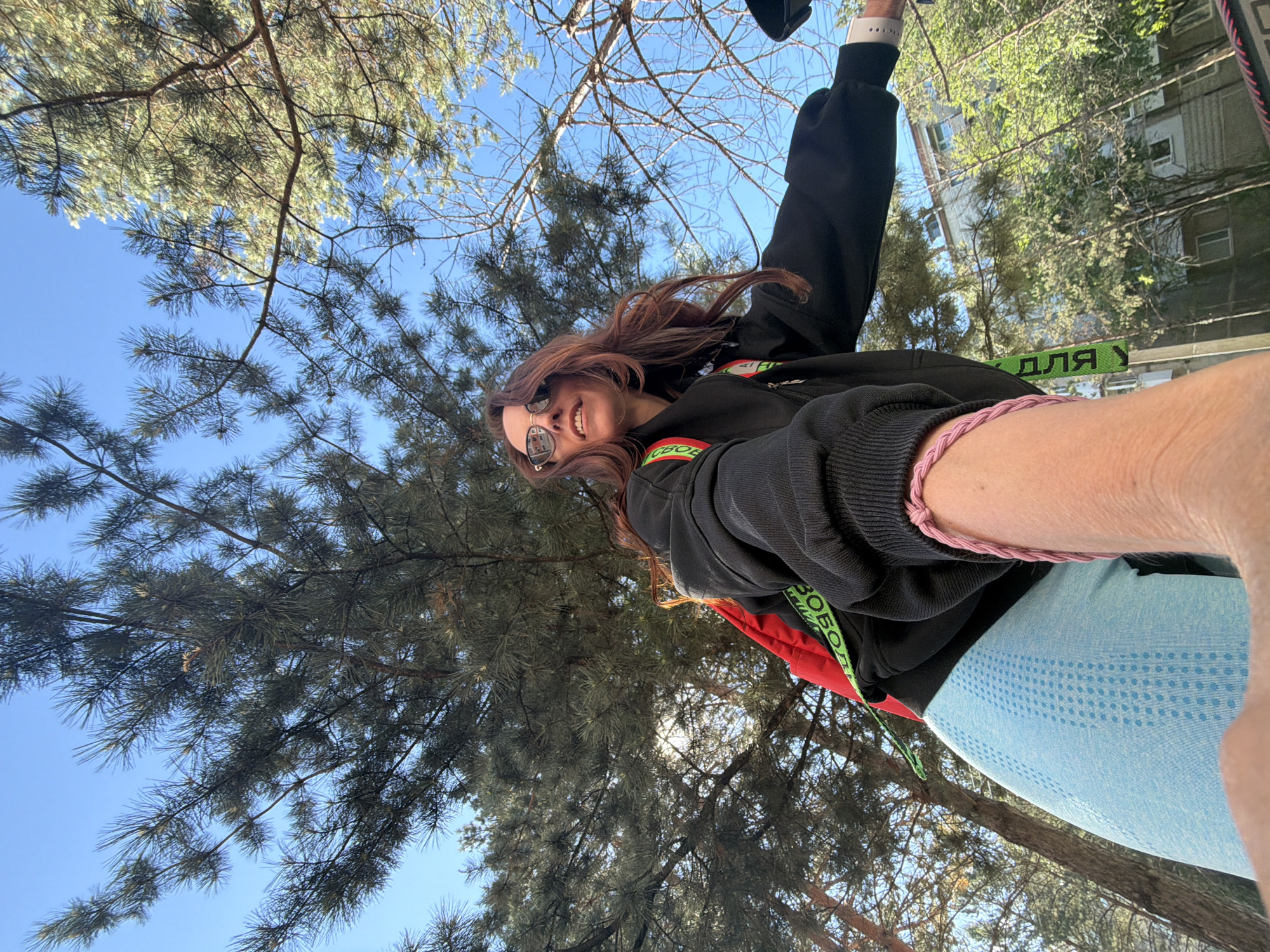 This image captures a low-angle selfie of a young woman outdoors in Birobidzhan, Russia, under a clear blue sky. The perspective is from below, looking up, giving an "upside-down" feel. The subject is a woman with long, reddish-brown hair, wearing sunglasses, a black hooded sweatshirt with colorful red and green details, and light blue patterned leggings. She is smiling widely, conveying happiness or contentment. On her right wrist, she wears a pink braided bracelet, and a white wristband (possibly a smartwatch) is visible on her left wrist.
The background is dominated by numerous pine or conifer trees with their branches and needles framing the bright, sunny sky. A multi-story residential building with windows is partially visible on the right side of the frame, peeking through the tree canopy, indicating an urban or suburban setting.
A green strap or ribbon is visible across the woman's shoulder, featuring Cyrillic text. Parts of the text read "ДЛЯ У" and "СВОБОД", strongly suggesting the word "СВОБОДА" (meaning "freedom") is present, possibly as "For Freedom". The photo appears to have been taken during daylight hours, given the bright sun and clear conditions. The overall scene depicts a casual, pleasant moment spent outdoors.