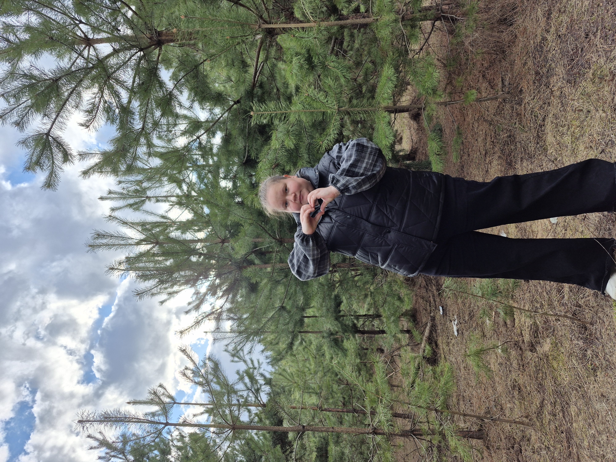 A photograph, taken in a forest or woodland area in Chaikovsky, Russia, captures a young person standing among pine trees. The individual, appearing to be a child or pre-teen with light-colored hair, is positioned centrally in the frame. They are dressed in a dark, possibly black, quilted vest worn over a grey and black plaid long-sleeved shirt, dark trousers, and light-colored shoes. The subject is actively posing, forming a heart shape with both hands in front of their chest, conveying a gesture of affection, love, or happiness.
The setting is an outdoor, natural environment dominated by numerous pine trees with vibrant green needles, forming a dense background. The ground beneath the trees is covered with dry, light brown grass and some patches of soil, indicating a season where ground vegetation is dormant, possibly late autumn or early spring. Above the tree canopy, the sky is visible, featuring a blend of clear blue patches and soft, white clouds, suggesting a partially cloudy daytime. The lighting is natural and bright, consistent with a midday or afternoon shot. No text is visible in the image. The scene portrays a casual, serene moment within a coniferous forest landscape.