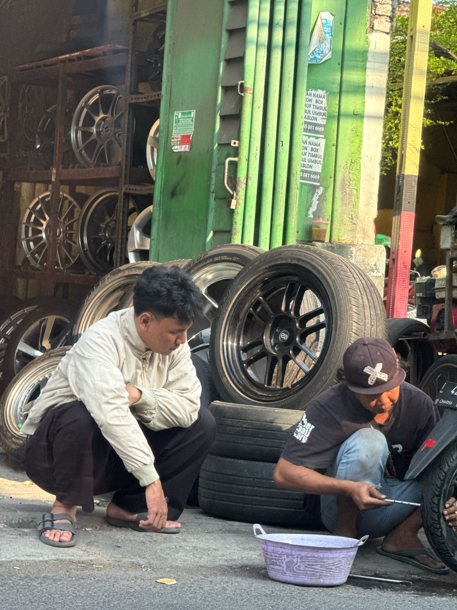 In Surabaya, Indonesia, on a sunny day, two men are crouched beside a collection of car tires and rims displayed outside a shop. The shop's exterior features a green metal roll-up door with peeling paint and a yellow and black striped pole. Racks of silver rims are visible in the background.
The man on the left, wearing a light-colored jacket and dark pants, is squatting with his arms crossed. He wears grey sandals.
The man on the right, dressed in a black t-shirt and jeans, is actively working on a tire. He wears a dark baseball cap and dark green flip-flops. He holds a tool and appears to be adjusting or repairing the tire, with a light purple basin sitting nearby. A motorcycle is partially visible on the right side of the frame, with its tire being worked on. The ground is paved with asphalt.