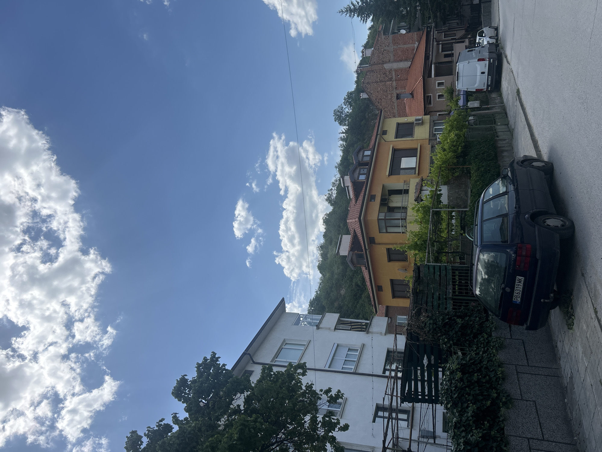 The image depicts a street scene in an urban area of Unknown, Unknown. A multi-story building with white facade is on the left side of the frame, partially obscured by trees. Adjacent to it is a yellow building with an orange tile roof, followed by a brick building with a similar roof. A green, vegetated slope rises in the background behind the buildings. A parked blue car occupies the foreground on the right side of the frame, near a light gray curb. A white van is parked further up the street, near another white car. The sky is predominantly blue with scattered white clouds. Overhead power lines are visible stretching across the sky.