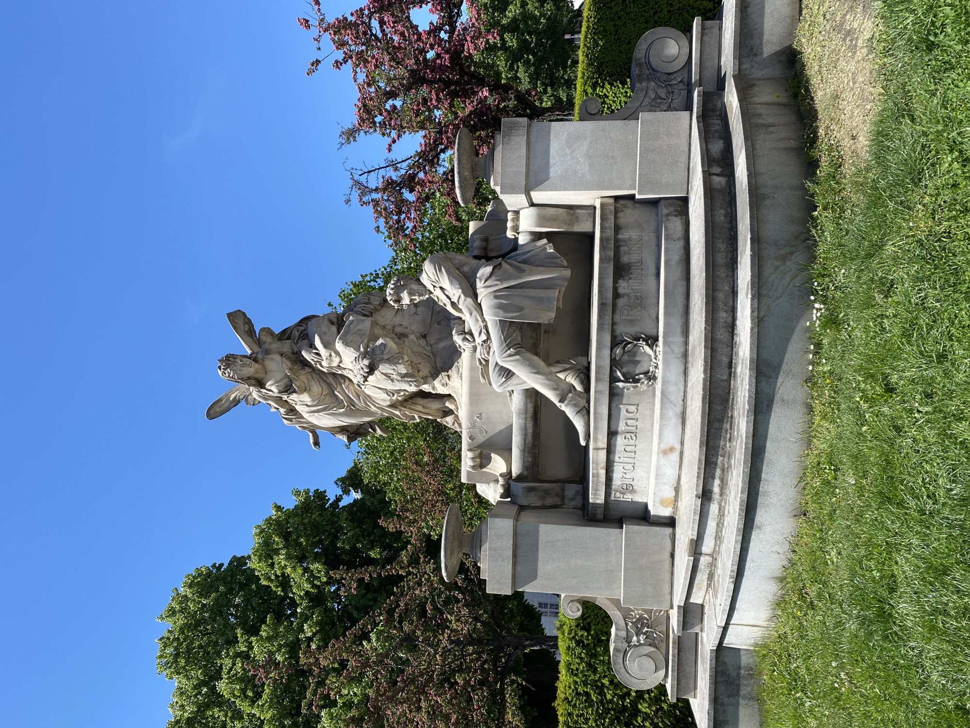 The photograph depicts the Raimund monument, located in Vienna, Austria. The monument, crafted from white stone, features a statue of a seated man (Ferdinand Raimund) with a sorrowful expression. An angel, positioned behind the seated man, appears to be supporting or consoling him. The monument also includes inscriptions with the names "Ferdinand" and "Raimund" and a wreath. The surroundings include green grass, trees with green and pink foliage, and a clear blue sky.