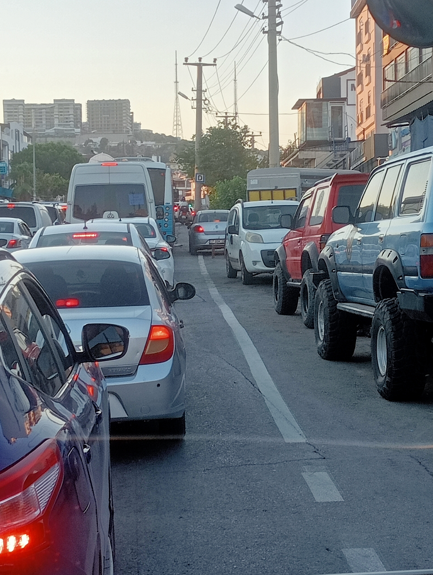 The image depicts a city street scene with heavy traffic. Several vehicles, including cars, vans, and what appears to be a bus, are stopped or moving slowly in a traffic jam. The vehicles are of various colors and models, with some prominent off-road vehicles with large tires visible. Buildings line the street, some of which appear to be under construction. Utility poles with wires are also present. The sky is overcast with a hazy quality, suggesting it might be early morning or late afternoon. The road surface shows some wear and tear. The perspective suggests the photo was taken from within a vehicle.
