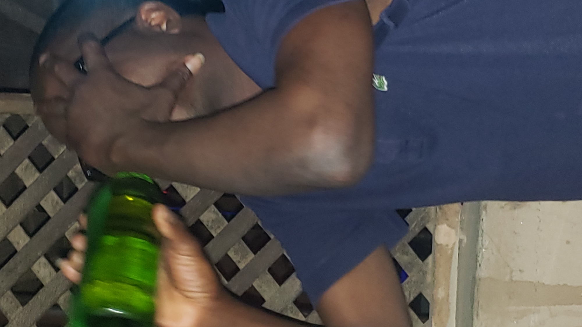 A person, likely male, is holding a green glass bottle. He is wearing a navy blue polo shirt. His right hand is obscuring part of his face. The background consists of a latticed structure. The location is Narayi, Nigeria.