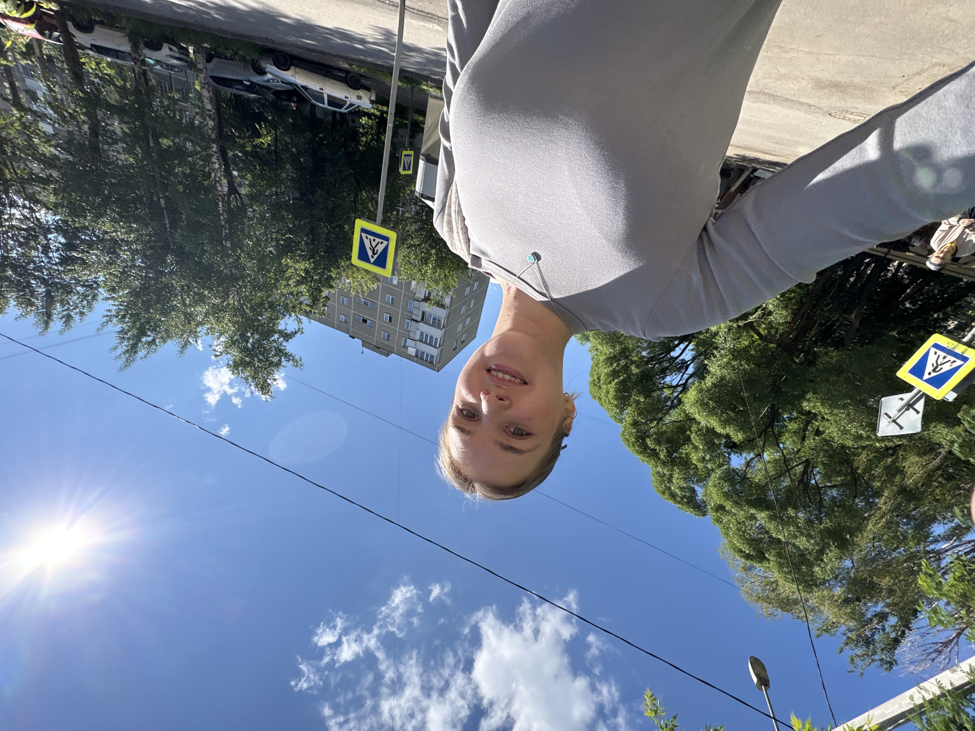 A selfie of a young woman stands against a backdrop of a sunny cityscape in Perm, Russia. The blue sky is partially obscured by fluffy white clouds and a bright sunburst. A pedestrian crossing sign with a blue and yellow symbol is visible, along with green trees lining the street. A multi-story building with several windows can be seen in the distance. The woman is wearing a gray top, and an earbud cord is visible. Utility wires stretch across the sky.