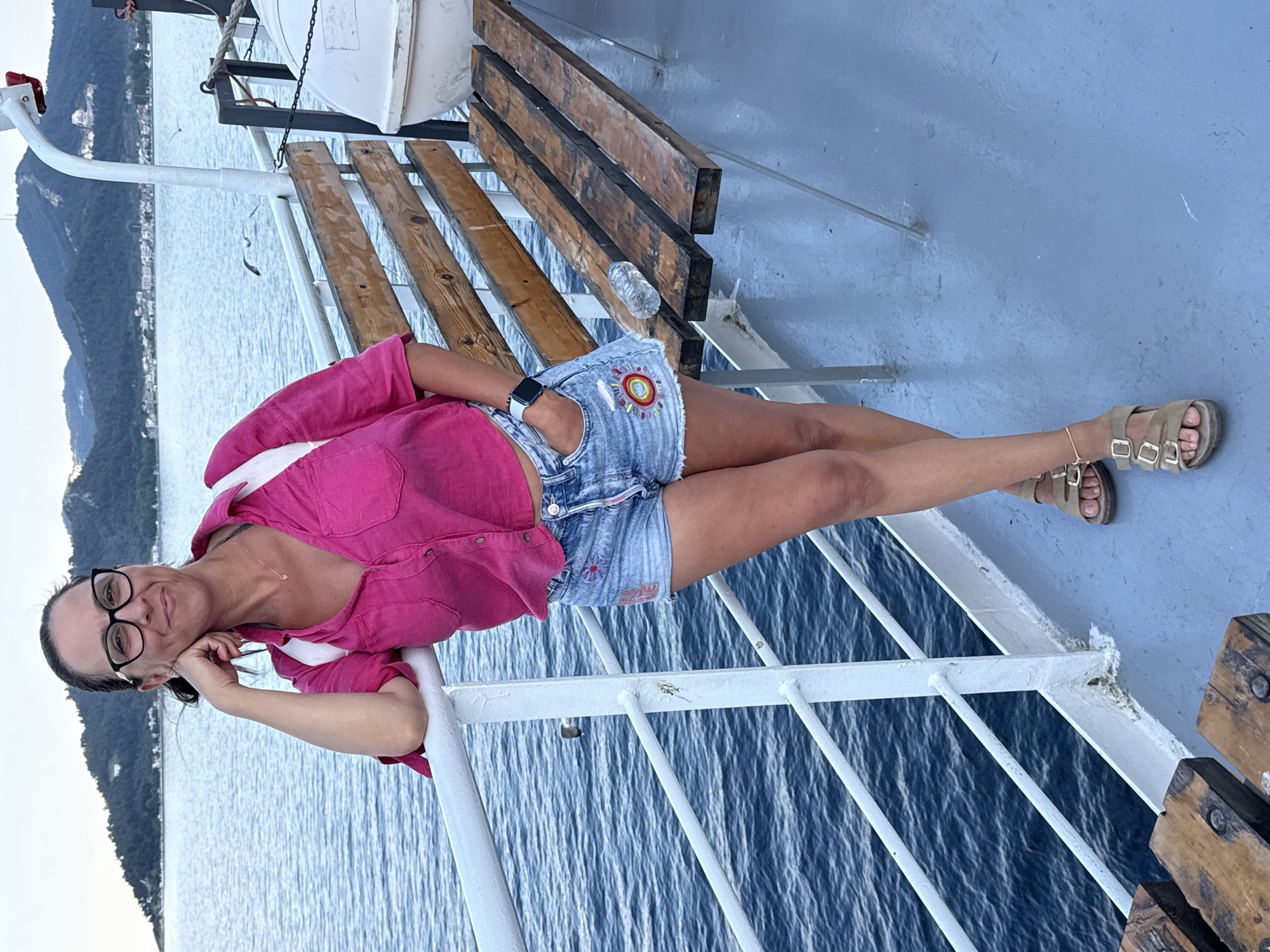 A woman wearing glasses, a pink button-up shirt, and denim shorts is standing on the deck of a ferry. She is leaning against the railing, with her left arm resting on it and her right hand in her pocket. The ferry is sailing on the water with a coastline and mountains in the background, which could be the coast of Thassos, Greece. The deck is painted a light blue, and there are wooden benches on the right side of the image. The water is a deep blue, and the sky is overcast.