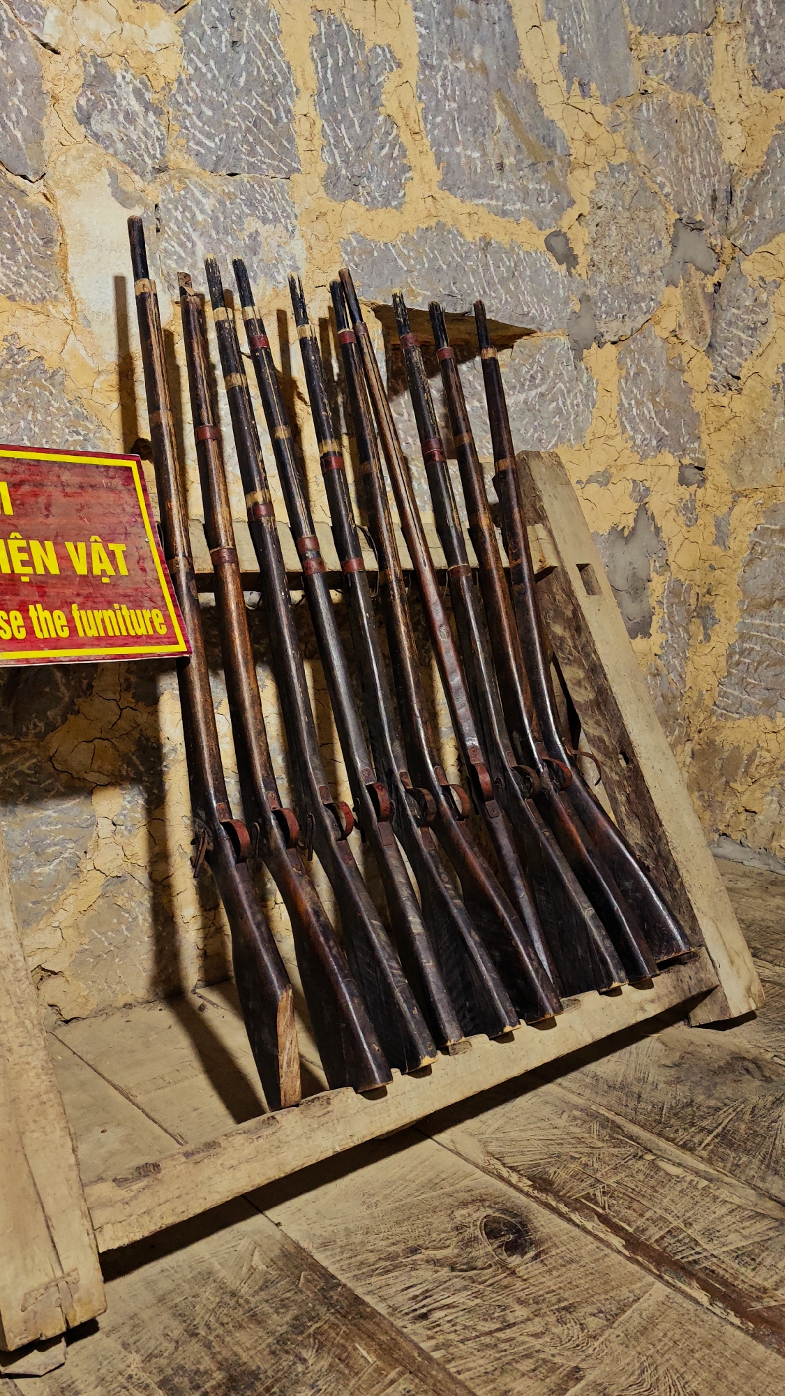 The image shows a collection of antique rifles displayed in a wooden rack against a textured wall. A sign in yellow and red lettering is partially visible on the left. The rack is situated on a wooden floor. This scene is located in the city of Sà Phìn Commune, Vietnam.