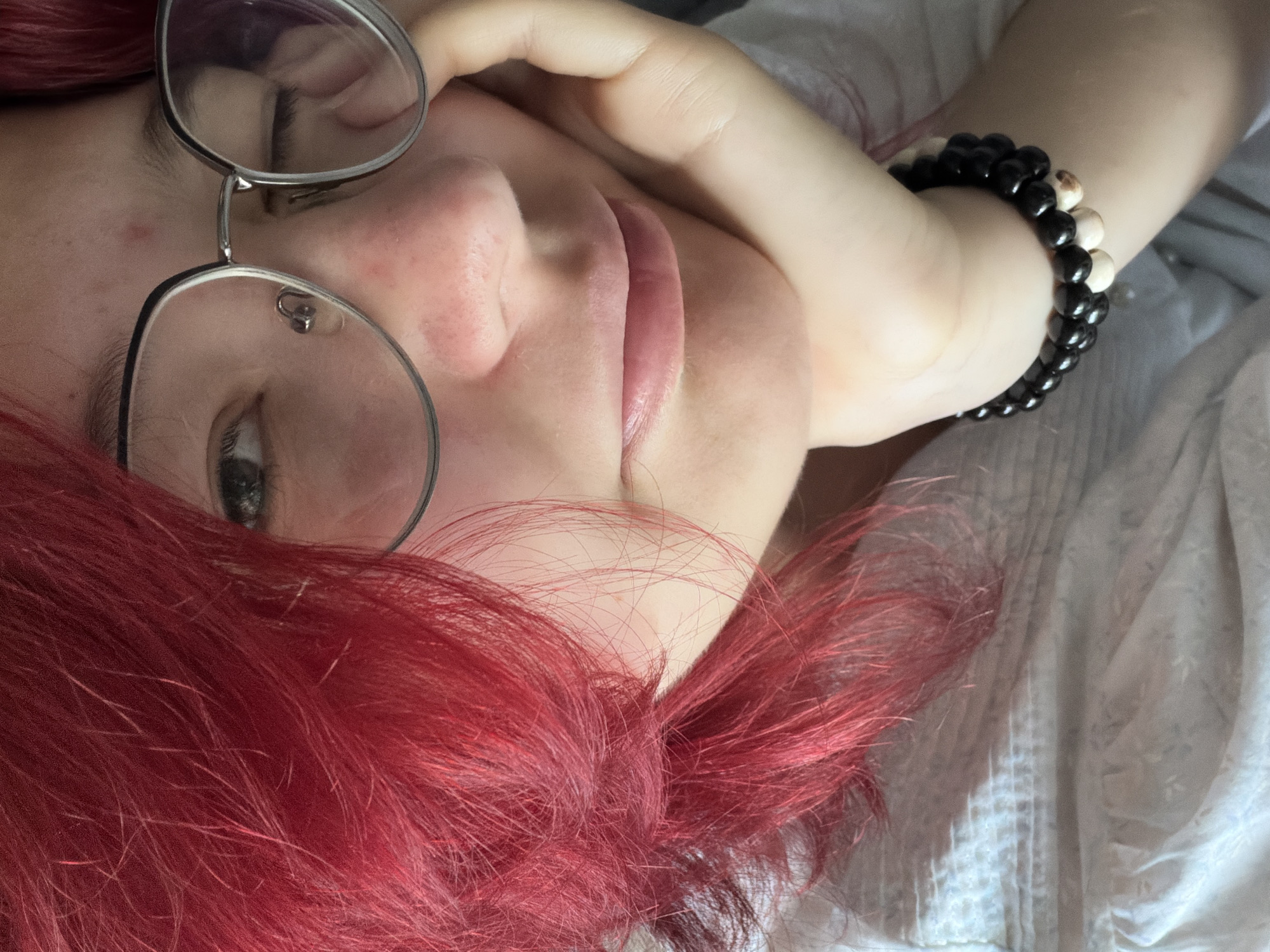 A close-up shot shows a young person with vibrant red hair, resting their head on their hand. They are wearing round, silver-rimmed glasses and a black and white beaded bracelet adorns their wrist. Their eyes are partially closed, and their lips are slightly parted. The background is a soft, textured white fabric. The image appears to be taken indoors, with no discernible view of the city of Chelyabinsk, Russia.