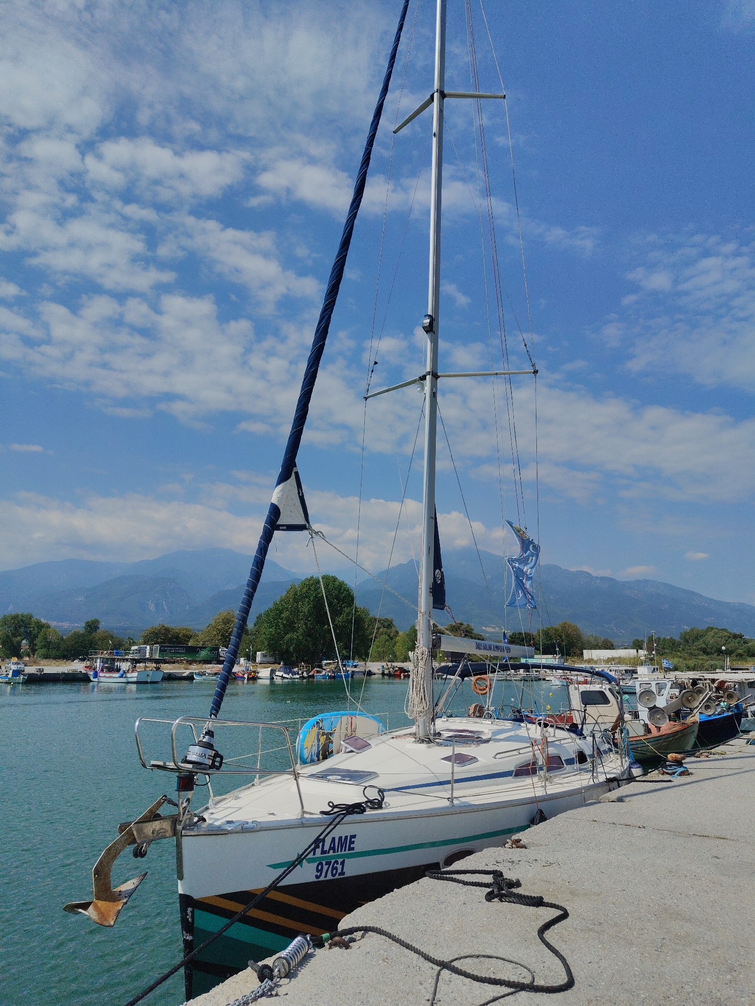 A sailboat named "FLAME 9761" is docked in a harbor under a blue sky with scattered clouds. The boat is white with green and yellow stripes along its hull. An anchor is extended from the bow, resting on the concrete pier. The mast is tall with several blue and white flags flying from it. Other boats are moored alongside the pier, including a fishing boat with nets and equipment. In the background, lush green trees and a range of mountains form a scenic backdrop. The scene appears to be a tranquil day in a Greek harbor, possibly Limani Litochorou, given the context. The time of day is likely daytime, with bright sunlight casting shadows. There are no people visible in the image. Visible text includes "FLAME 9761" on the boat's hull and "DAILY SAILING XPERIENCE K-128" on a banner on the boat.