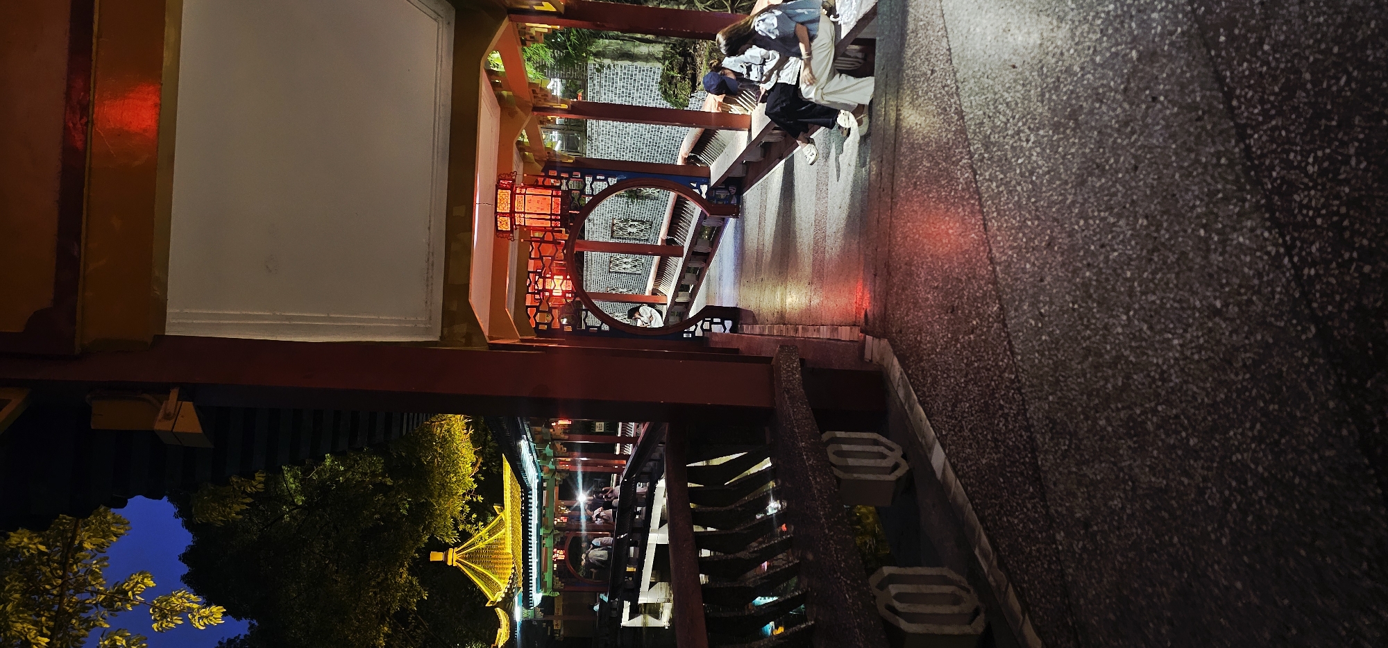 The image depicts a scene in Yuzhong District, China, at night. The foreground shows a pedestrian walkway with a textured surface. An ornate, traditional Chinese structure frames the left side of the image, featuring red pillars and decorative elements. Lanterns are visible within the structure, emitting a red glow. A couple is visible walking along the walkway, with one person wearing a blue shirt and white pants, and the other wearing black pants and a white top. Further in the background, the illuminated roof of a traditional Chinese building can be seen through an opening in the structure. Trees frame the left side of the view of the building, with a dark blue sky visible above.