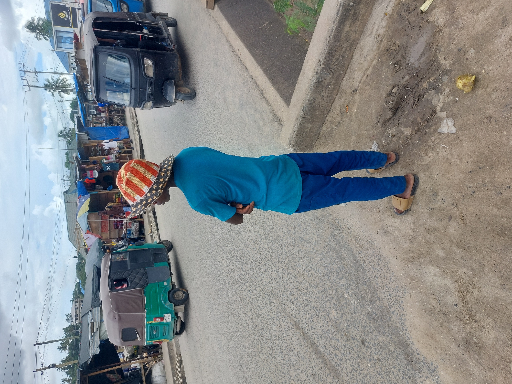 A person stands on a paved street in Dar es-Salaam, Tanzania. The individual wears a blue shirt, blue pants, a hat with an American flag-inspired design, and sandals. To the left, there are three-wheeled vehicles (tuk-tuks) along the street, with market stalls and buildings visible in the background. To the right of the person is a concrete curb and a patch of dirt with some debris. The weather appears to be partly cloudy.
