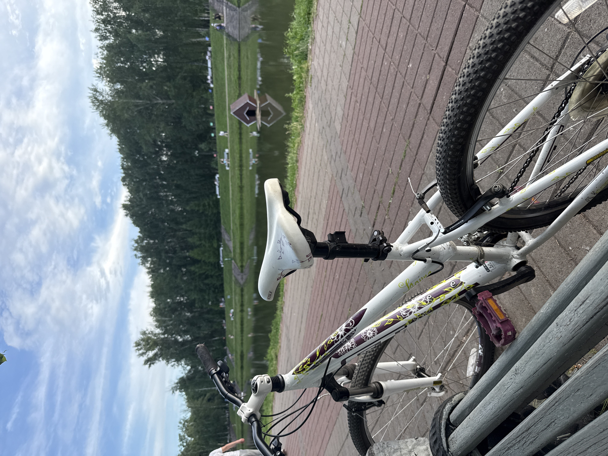 This media file shows a white bicycle parked next to a wooden bench. The bicycle is oriented with its wheels facing left. The scene appears to be outdoors, possibly at a park or recreational area. In the background, there is a body of water with a wooden structure in it, and beyond that, a treeline. The sky is cloudy and bright, suggesting it is daytime, likely late morning or afternoon. The weather appears to be mild.
The bicycle itself is white with purple and green floral decals on the frame. It has a white seat, black handlebars, and a black tire. The front brake is visible and appears to be a Promax model. The rear wheel is partially obscured by the bench. A purple pedal is visible on the right side of the bike.
The background includes a paved walkway made of reddish-brown bricks. The water is calm and reflects the sky and the wooden structure. There are several small figures of people visible in the distance, near the water and the treeline, suggesting the presence of other visitors enjoying the area. One individual's legs are visible in the lower left corner of the frame, indicating someone is sitting on the bench.
There is some text visible on the bicycle frame. On the down tube, above the pedal, "SAHARA" is printed in a green script. Above the rear wheel, a yellow sticker with black text that is partially visible and appears to say "WARR..." and another sticker below it that is unreadable.
The overall impression is one of a pleasant, casual outdoor setting, possibly a park with a lake or pond. The scene does not convey any specific strong emotions, but rather a sense of tranquility and outdoor recreation. The location is identified as Salavat, Russia.