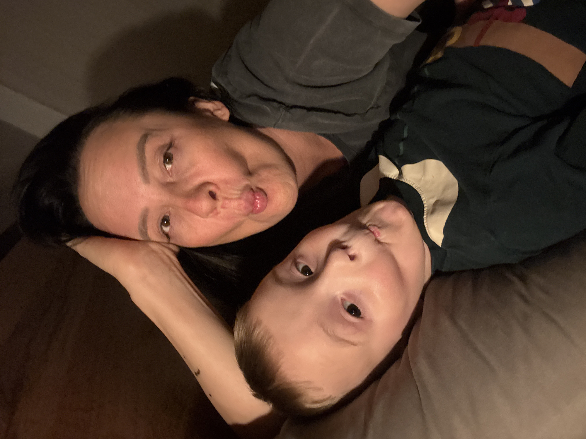 An overhead shot shows a woman and a child lying down, both with pursed lips. The woman is wearing a dark gray t-shirt and has dark hair. The child is wearing a dark green t-shirt with a light-colored collar. They appear to be lying on a brown couch or bed. The location is unknown.