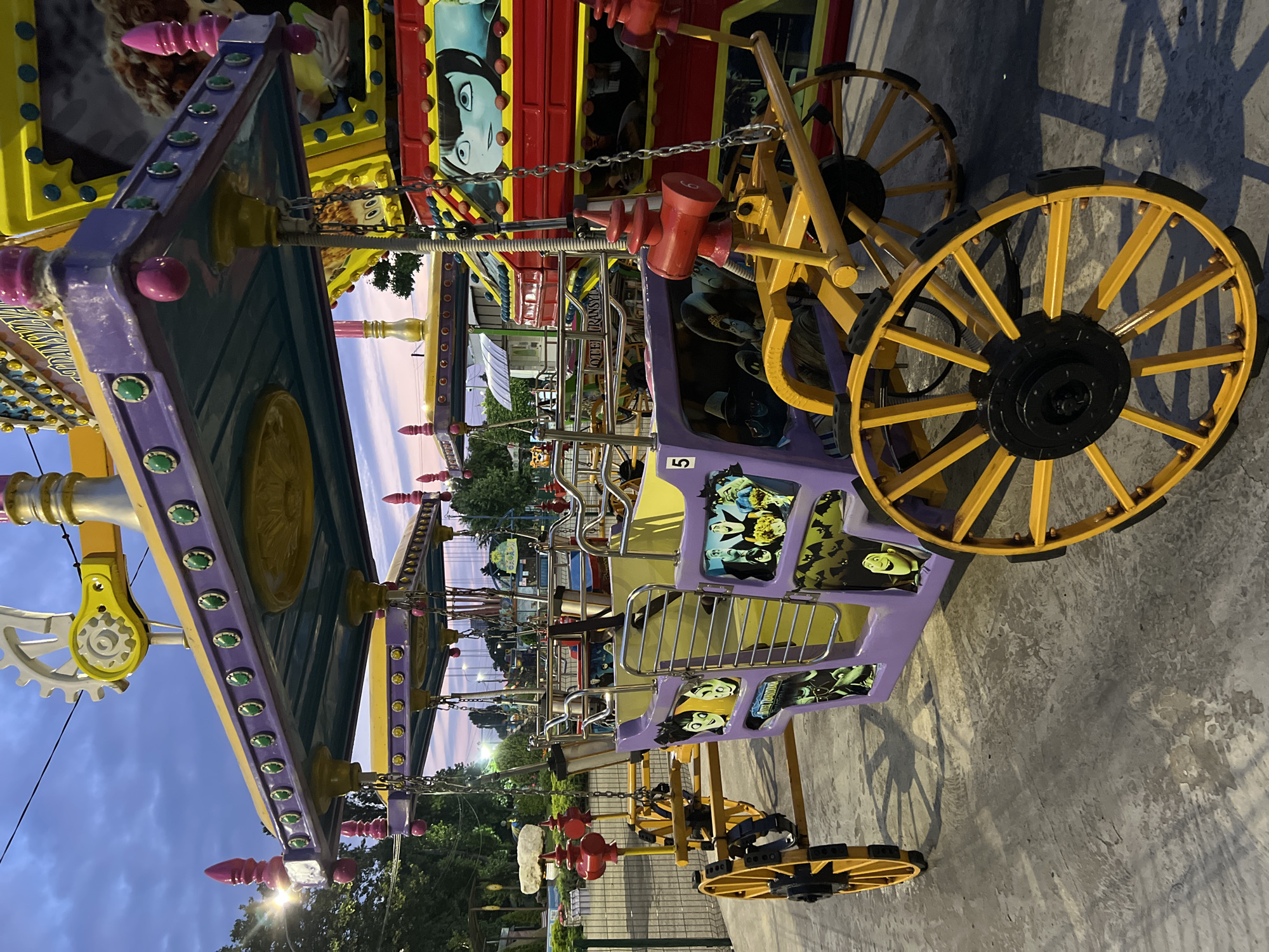 This image shows a brightly colored amusement park ride at dusk. The ride appears to be a themed carriage with characters from the movie "Hotel Transylvania" painted on the side. The carriage is purple and yellow with large yellow spoked wheels. The ride is suspended by chains from a larger structure, also decorated with colorful elements and lights. In the background, other amusement park rides are visible, including a Ferris wheel and a roller coaster. The sky is a gradient of purple and orange, indicating sunset. The ground is a paved surface. The location is not specified as Krasnodar, Russia in the image or provided context.