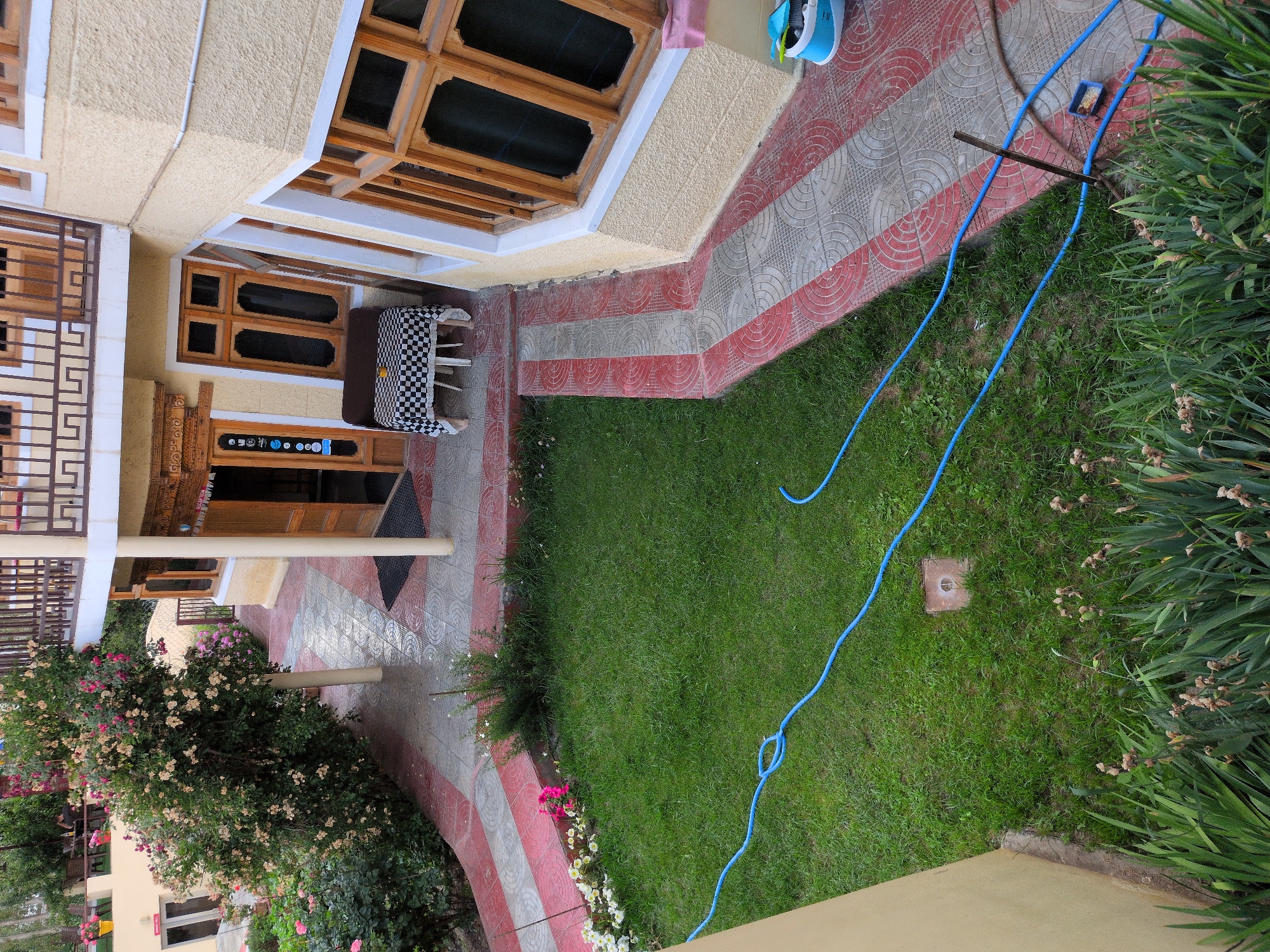 This image depicts the exterior of a building in Leh, India, likely a guesthouse or hotel. The building has a pale yellow stucco exterior with wooden-framed windows. A balcony with wooden railings is visible on the upper floor. The foreground shows a well-maintained green lawn with a blue garden hose laid across it. A square, concrete cover is partially visible in the grass. A paved pathway with red and gray geometric tiles leads to the building entrance. Beside the pathway, a flowering bush with pink and white flowers is in bloom. A dark brown sofa is placed outside the entrance under a checkered tablecloth. The entrance itself is a wooden door with ornate carvings. The scene suggests a peaceful and well-kept outdoor space.