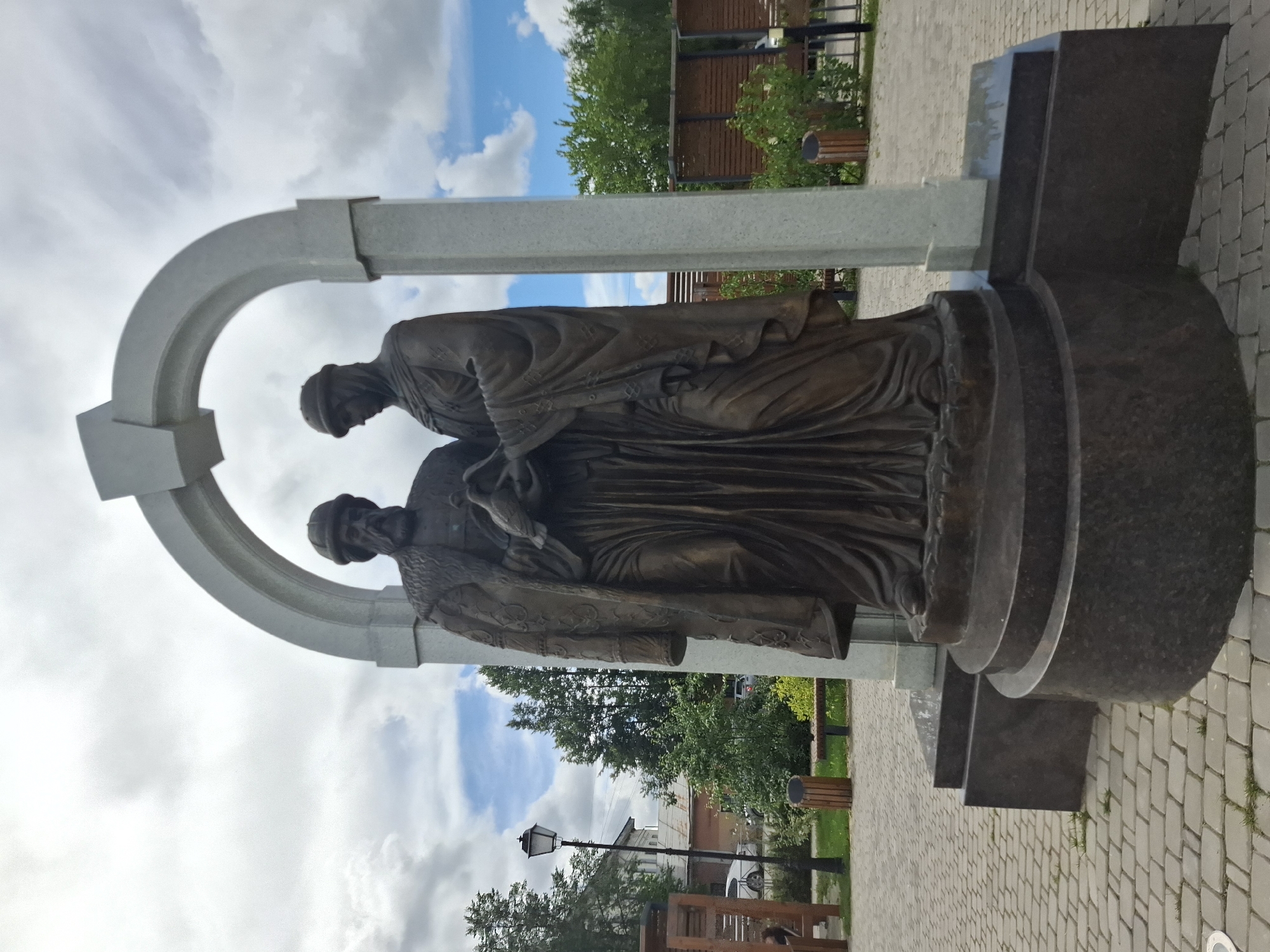 The image shows a bronze statue of two figures standing within a light-colored archway. The figures are dressed in historical clothing. The male figure on the left has a beard and wears a fur-lined coat and a cap. The female figure on the right wears a head covering and a long robe. They are both holding a dove or pigeon. The statue is on a dark granite pedestal. The scene is outdoors, with a paved walkway in the foreground. In the background, there are trees, buildings, a lamppost, benches, and trash cans. The sky is cloudy. This is a monument to St. Peter and St. Paul in Kungur, Russia.