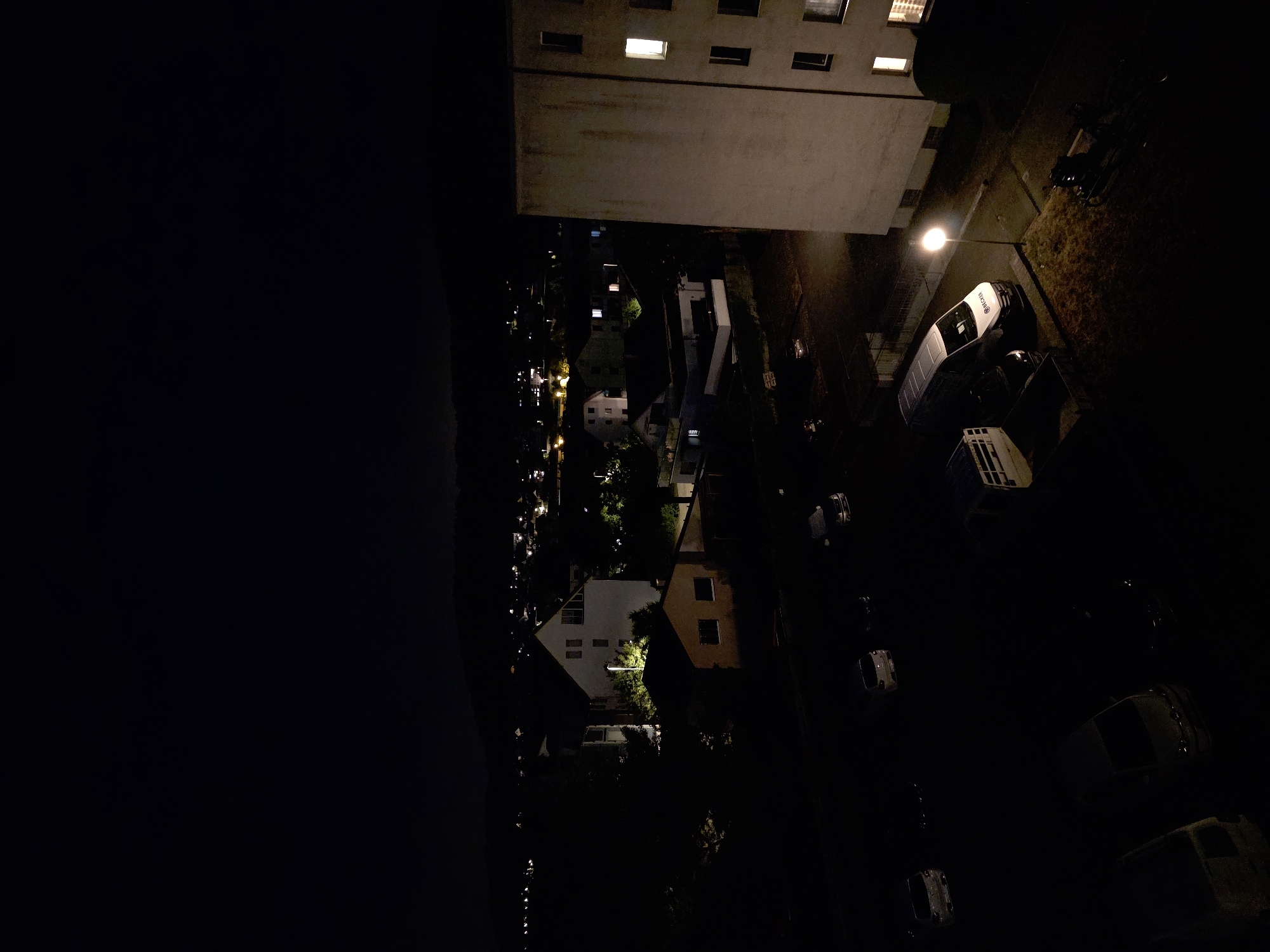 The image shows an aerial view of a residential area at night in Stein, Switzerland. The foreground is dominated by a large, light-colored apartment building on the right, with several windows illuminated. In front of the building, a street light illuminates a patch of grass and pavement. Several vehicles are parked on the street and in a lot, including a white van with "BECKER" written on its side.
In the middle ground, there are several houses and smaller buildings, some with lights on. Trees and foliage are visible, casting shadows in the darkness. The background shows a distant view of more illuminated buildings and houses, suggesting a broader urban or suburban landscape. The sky is dark, with a faint horizon line indicating a distant landscape. The overall scene depicts a quiet night in a residential neighborhood.