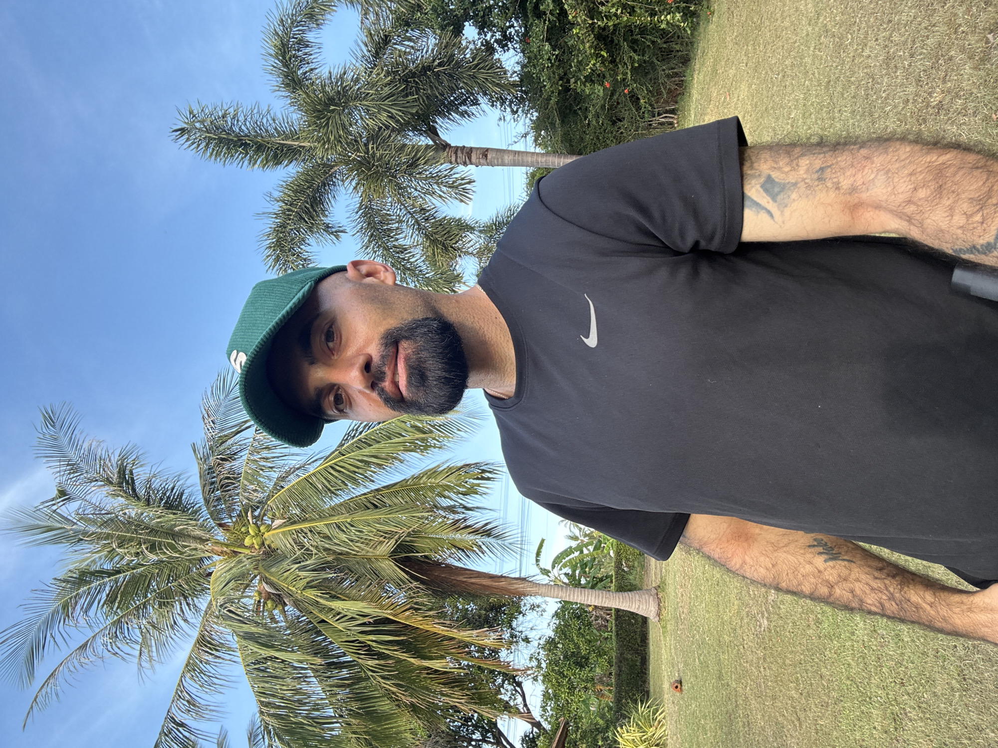 A man with a beard and a green cap stands in a grassy area with palm trees and lush vegetation in the background. He is wearing a black t-shirt with a grey Nike logo. He has tattoos on his right arm. The sky is clear and blue. The scene is likely outdoors in a tropical or subtropical location, consistent with Ko Samui District, Thailand.