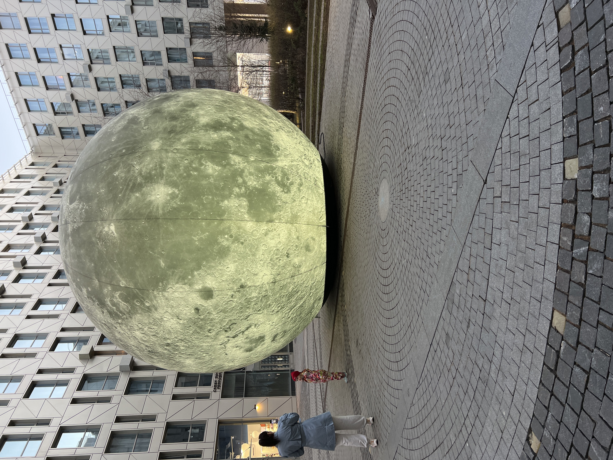 The image shows an outdoor scene in Saint Petersburg, Russia, featuring a large spherical sculpture resembling the moon. The sculpture is illuminated, displaying a detailed surface texture of craters and markings. The sculpture is positioned on a paved area made of light gray bricks arranged in concentric circles and squares that meets the surface of a modern building with repetitive window patterns. Two individuals are present: a woman in a light blue jacket and beige pants is looking at the sculpture, and nearby is a child wearing colorful clothing and a red hat. The image captures the interaction between the sculpture, the architecture, and the pedestrians in the urban environment.