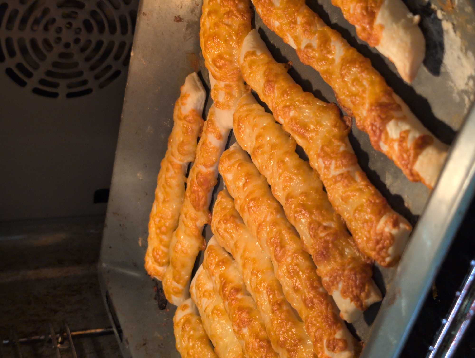This is a close-up, overhead shot of golden-brown cheese-covered breadsticks baking in a dark oven. The breadsticks are arranged in a tray, with some overlapping. They appear to be twisted and have a slightly crispy texture, with melted cheese clinging to their surfaces. The background shows the interior of the oven, including a fan grill on the left and oven racks on the right. The lighting is dim, typical of an oven's interior during baking. The overall impression is of freshly baked, savory snacks.