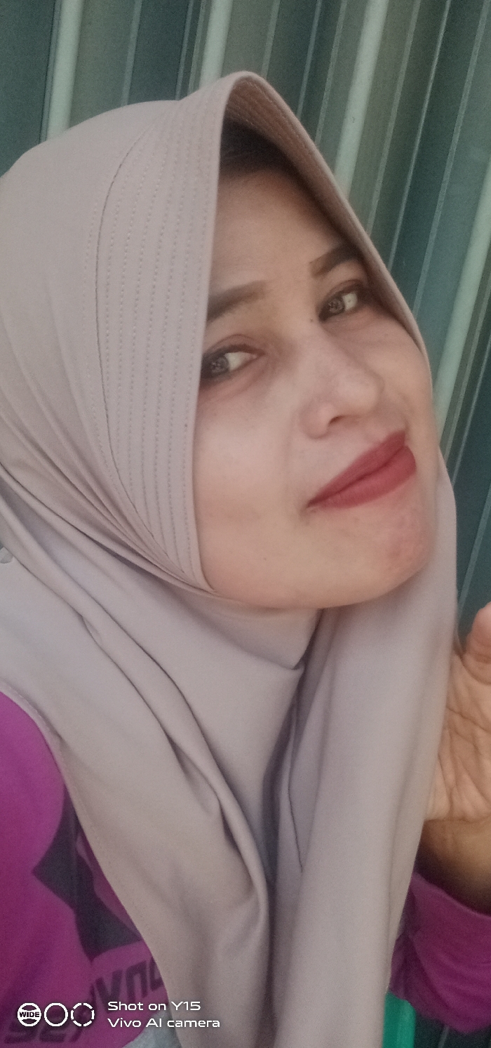 The image presents a close-up, chest-up portrait of an individual, presumably female, in what is identified as Gending, Indonesia. The person's head is slightly tilted to their right, and they are looking towards the camera with a subtle smile. They are wearing a light taupe or beige hijab, which covers their head and neck, displaying a vertical stitched or ribbed pattern along the forehead section. Dark eyebrows, dark eyes, and dark red lipstick are visible on the face. A magenta-colored garment is visible beneath the hijab on the left side of the frame, and a partial view of a light-skinned hand is present on the lower right. The background features vertical stripes in varying shades of teal and dark green, suggesting a patterned wall or a closed blind. A watermark in the lower left indicates the image was "Shot on Y15 Vivo AI camera."