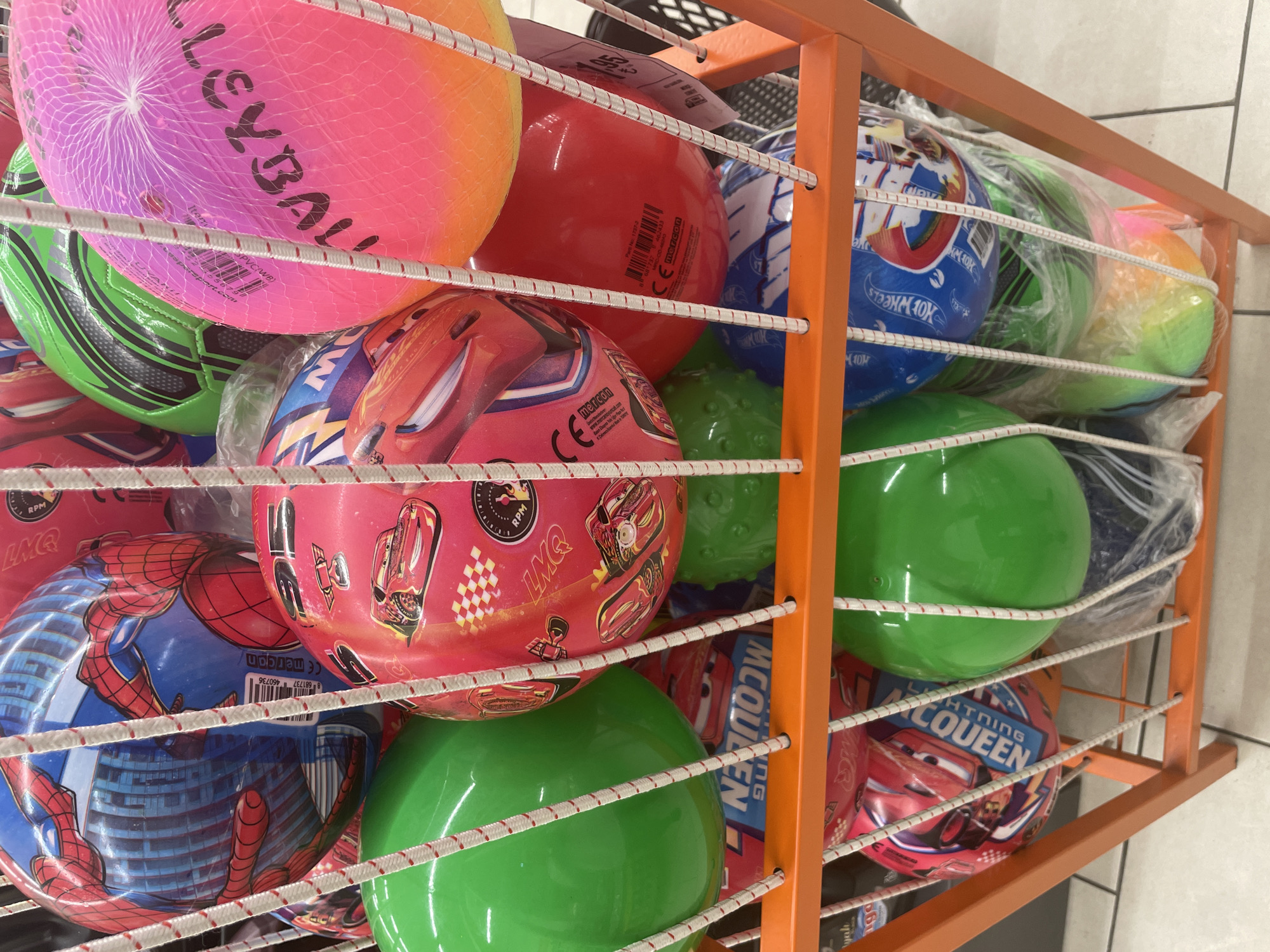 The image shows a close-up, eye-level, vertical shot of a display rack filled with colorful inflatable balls. The rack is made of orange metal bars, and the balls are arranged in rows, separated by white elastic cords. The balls vary in color and design. In the top row, there's a large pink and orange volleyball net ball, a solid red ball, and a green ball. Below that, a green soccer ball with black accents is visible, next to a collection of balls with popular cartoon characters. One ball prominently features Spider-Man, with his red and blue costume and buildings in the background. Another ball is adorned with images of Lightning McQueen from the movie Cars, with the text "LIGHTNING MCQUEEN" clearly visible. Some balls are partially enclosed in clear plastic packaging.
The scene appears to be an indoor retail environment, likely a toy store or a department store section dedicated to sporting goods or children's toys, given the display method and the types of products. There are no people visible, and the lighting suggests an indoor setting. The focus is entirely on the product display.
Visible text includes brand names like "mercan," product-related terms like "VOLLEYBALL," "PVC(WB)," and character names like "SPIDER-MAN" and "LIGHTNING MCQUEEN." There are also product codes and potentially website addresses, such as "spirsport.com" and "www.летсапоулсай.com" which appears to be a Cyrillic transliteration of a Turkish website. The overall impression is one of a vibrant and diverse collection of play balls for children.