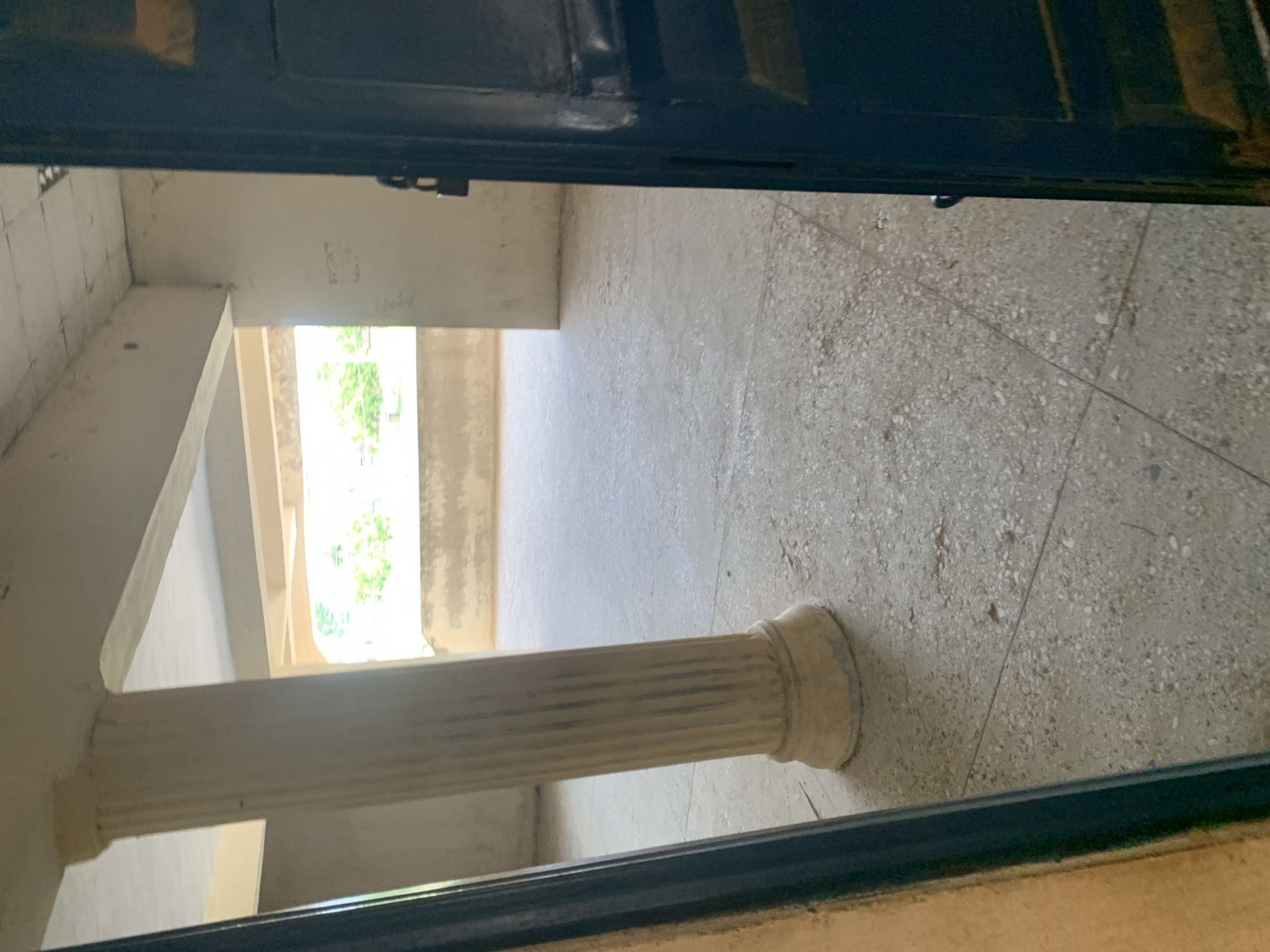 The image depicts an interior view of an open doorway, looking out into a large, unfinished or under-construction space. The scene is dominated by concrete textures. A fluted column, likely made of concrete or plaster, is prominently featured in the foreground, supporting a concrete beam. Beyond the column, the space opens up to reveal more concrete floors and walls. A large opening, possibly a window or another doorway, allows natural light to enter, illuminating the area and providing a view of greenery outside, suggesting the presence of trees or vegetation. The time of day appears to be daytime, with bright light filtering in. The overall impression is one of an unfinished building, possibly in a tropical or sub-tropical location given the greenery. There are no people visible in the image. Some faint markings or graffiti are present on the wall in the background, but they are illegible. The perspective is from within a dark doorway, looking out into a brighter, open space.