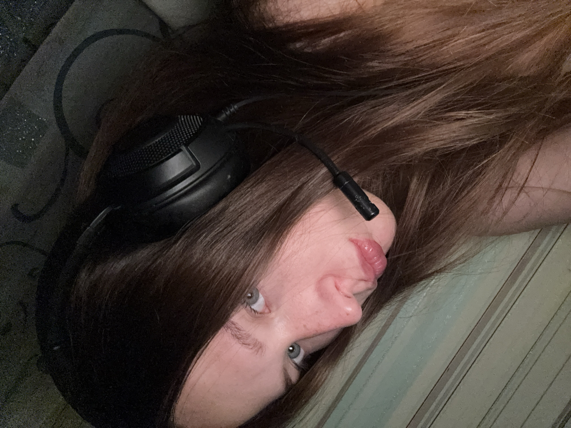 A young person with long, brown hair and blue eyes is pictured in a close-up, profile view, wearing black headphones with a microphone. They are looking towards the viewer with their lips pursed. The background is a striped pattern in muted green and beige tones. The image appears to be a selfie taken indoors. There is no information to suggest this image is located in Chaikovsky, Russia.